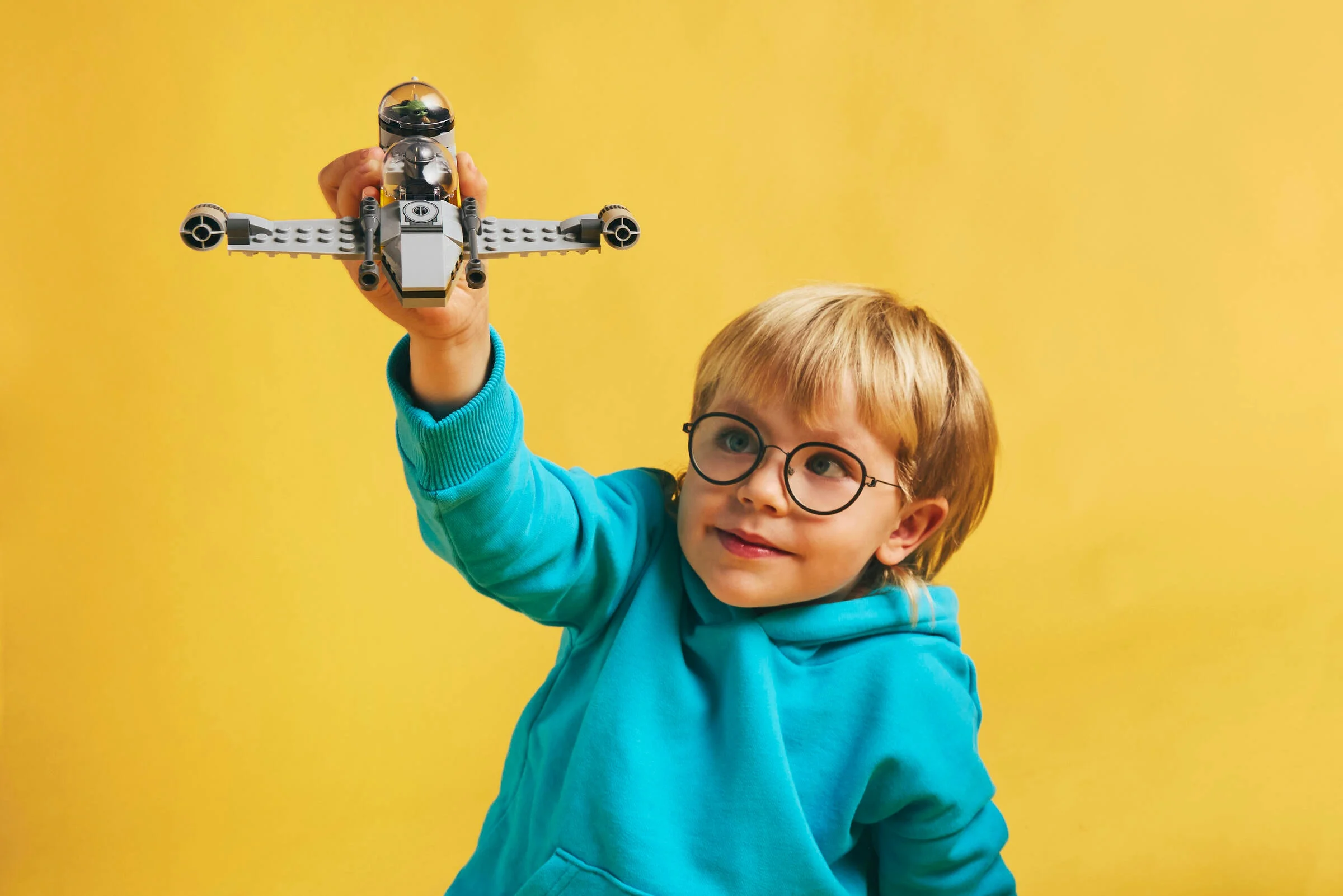 Copil se joacă cu N-1 Starfighter LEGO, zâmbind. Fundal galben, atmosferă jucăușă.