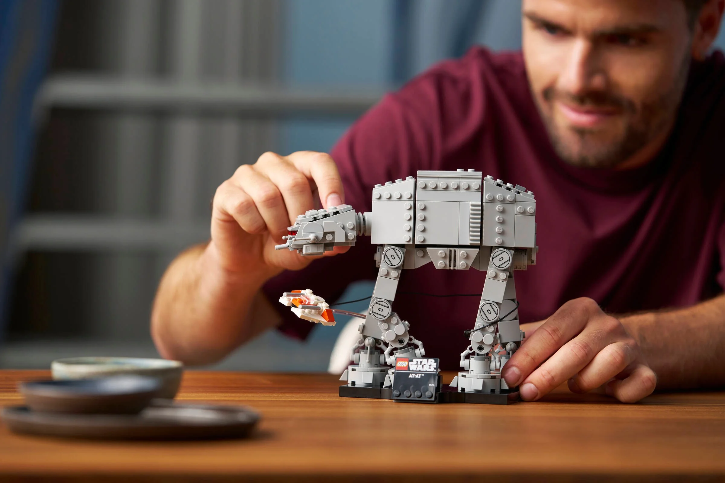 Bărbat construind AT-AT LEGO. Model gri, detaliat, pe o masă de lemn.
