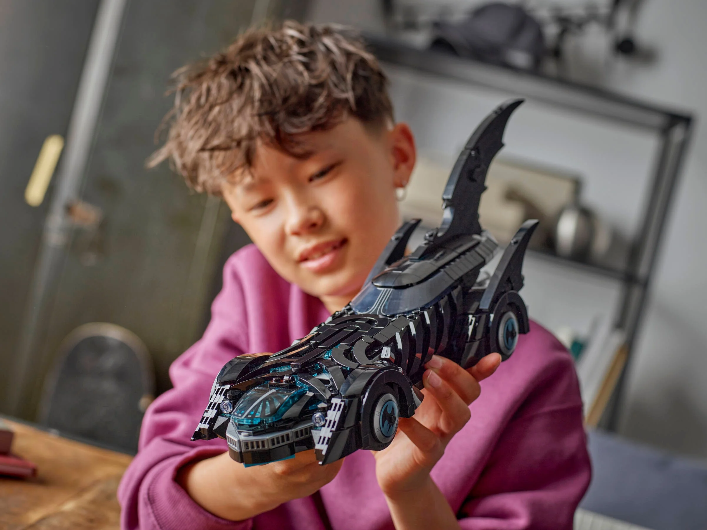 Băiat ține în mâini Batmobile-ul LEGO, zâmbind admirativ.