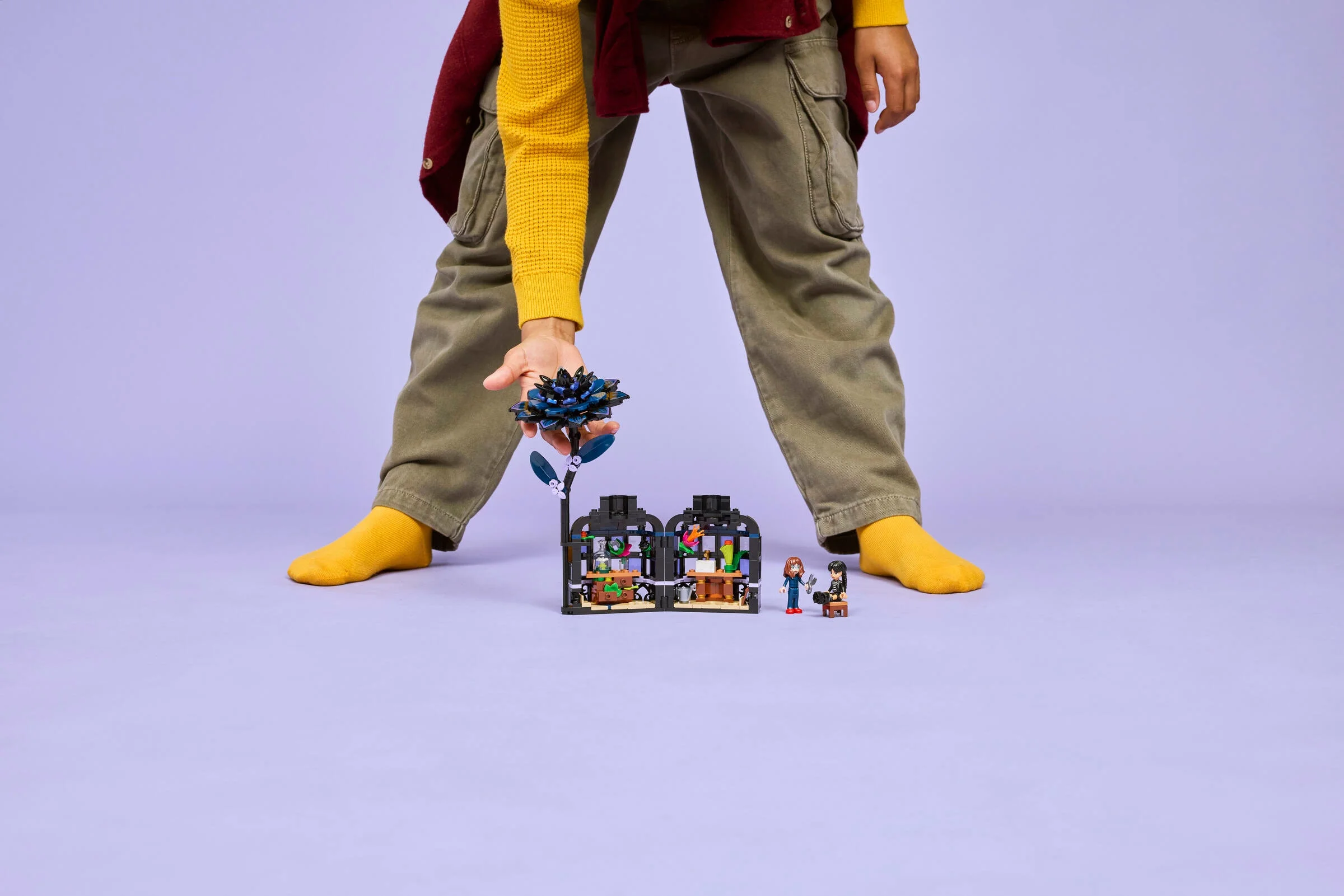 Copil ținând floarea Dahlia Neagră LEGO lângă serele cu minifigurine Wednesday.