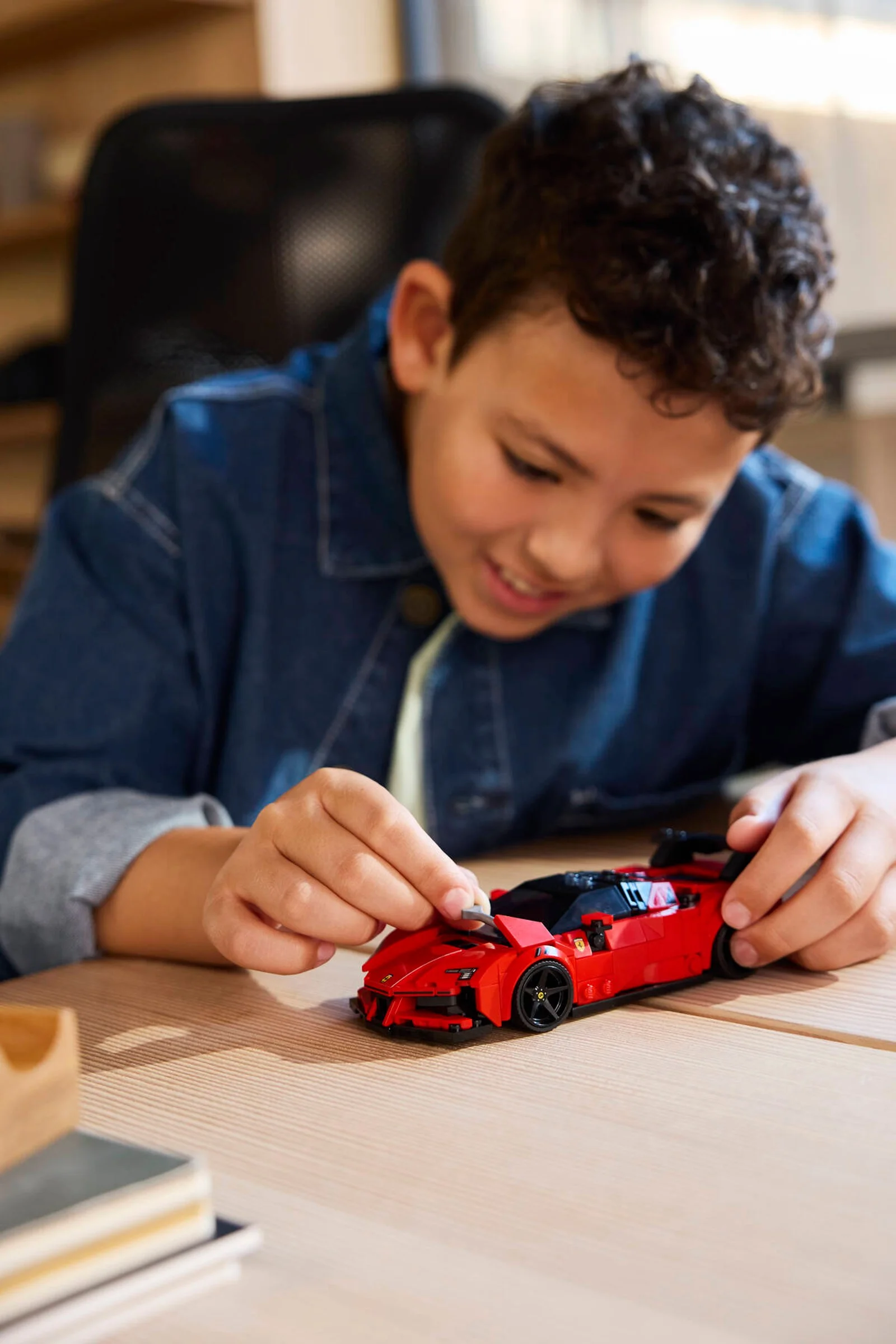 Un băiat zâmbind construiește modelul LEGO Ferrari SF90 XX Stradale roșu pe o masă de lemn.