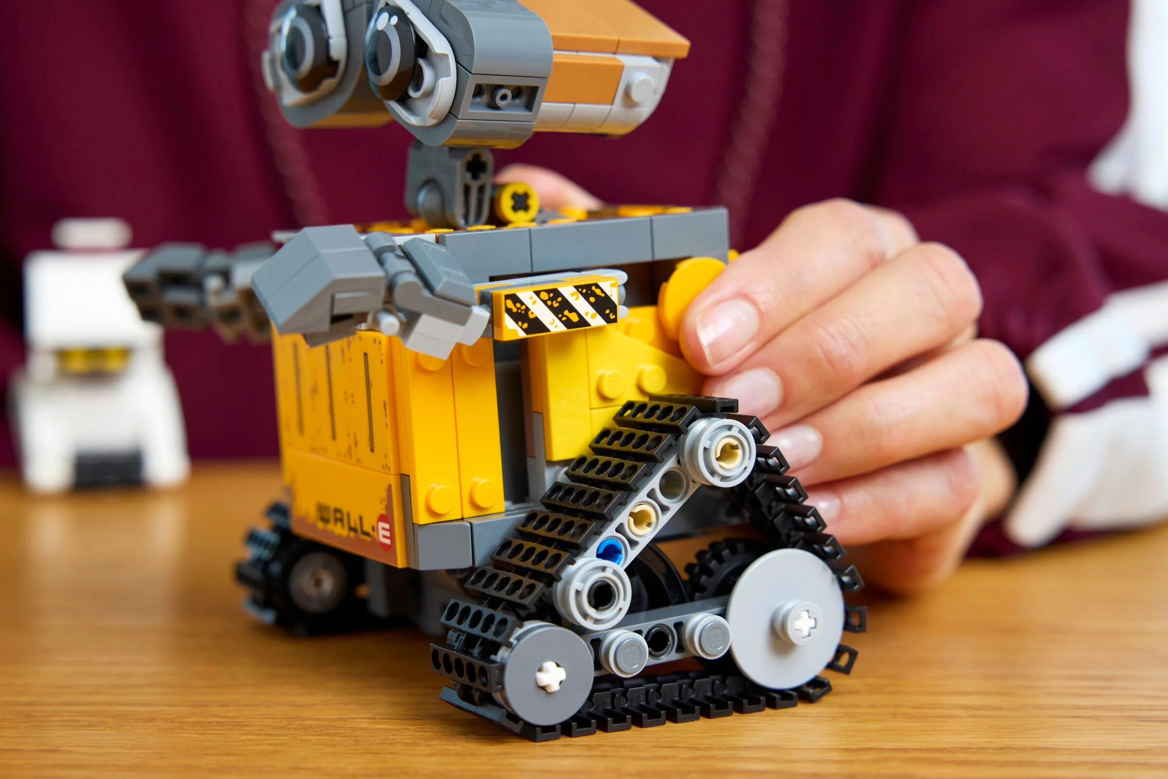 Prim-plan cu WALL-E LEGO, mâna unei persoane ajustându-i șenilele. EVE se vede estompată în fundal.