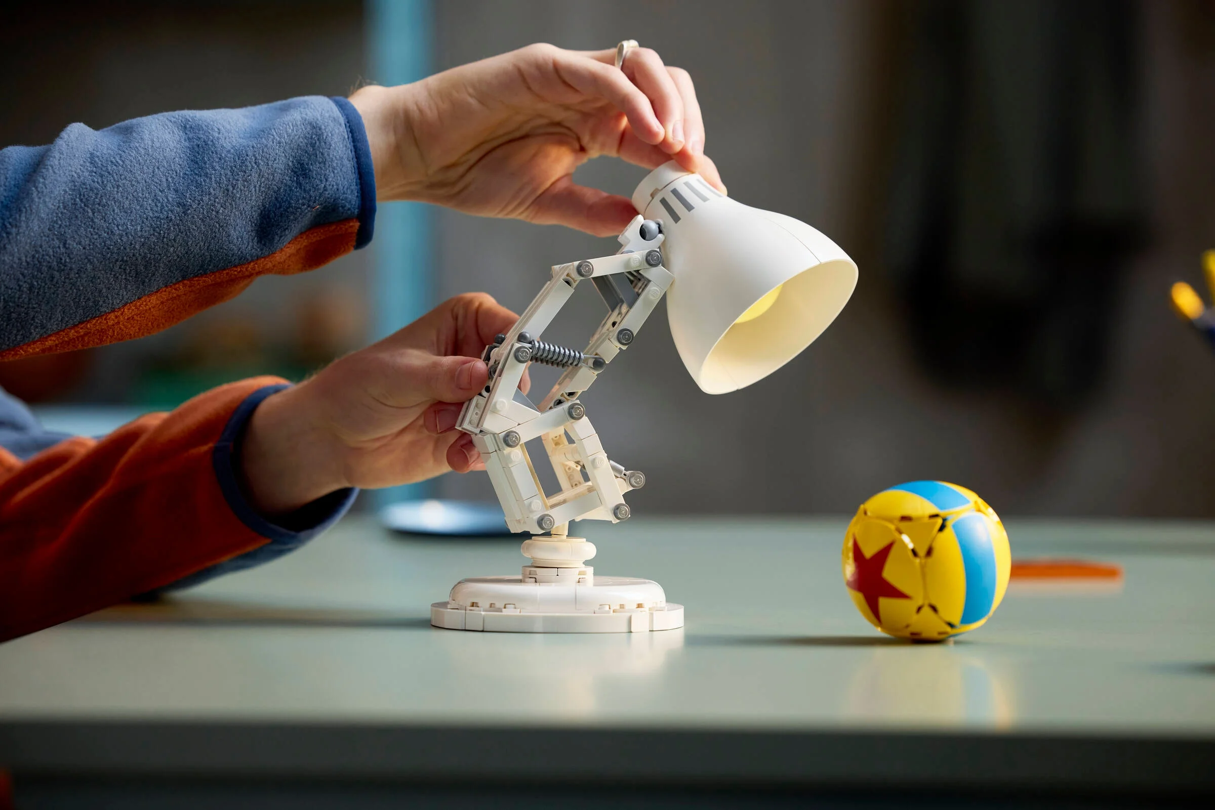 Modelul LEGO Luxo Jr. este ajustat de mâini, cu mingea Pixar lângă.