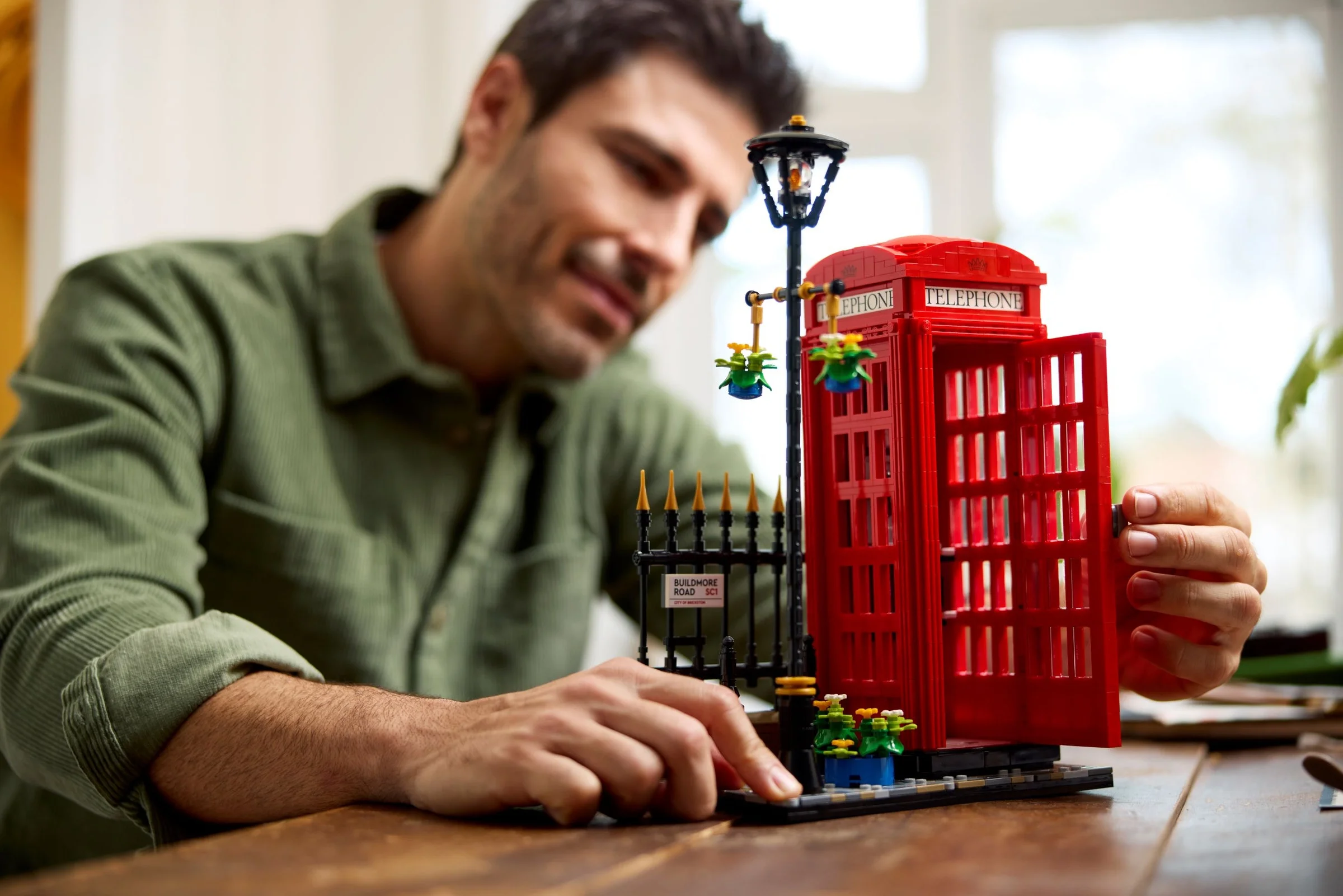 Bărbat interacționând cu cabina telefonică roșie LEGO, o deschizând. Un detaliu al scenei construite.