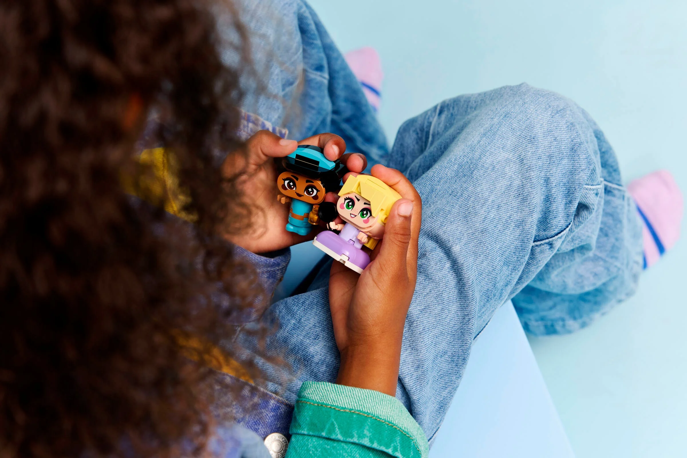 O fetiță ține în mână mini-figurinele Lego Jasmine și Rapunzel, stând pe un scaun.