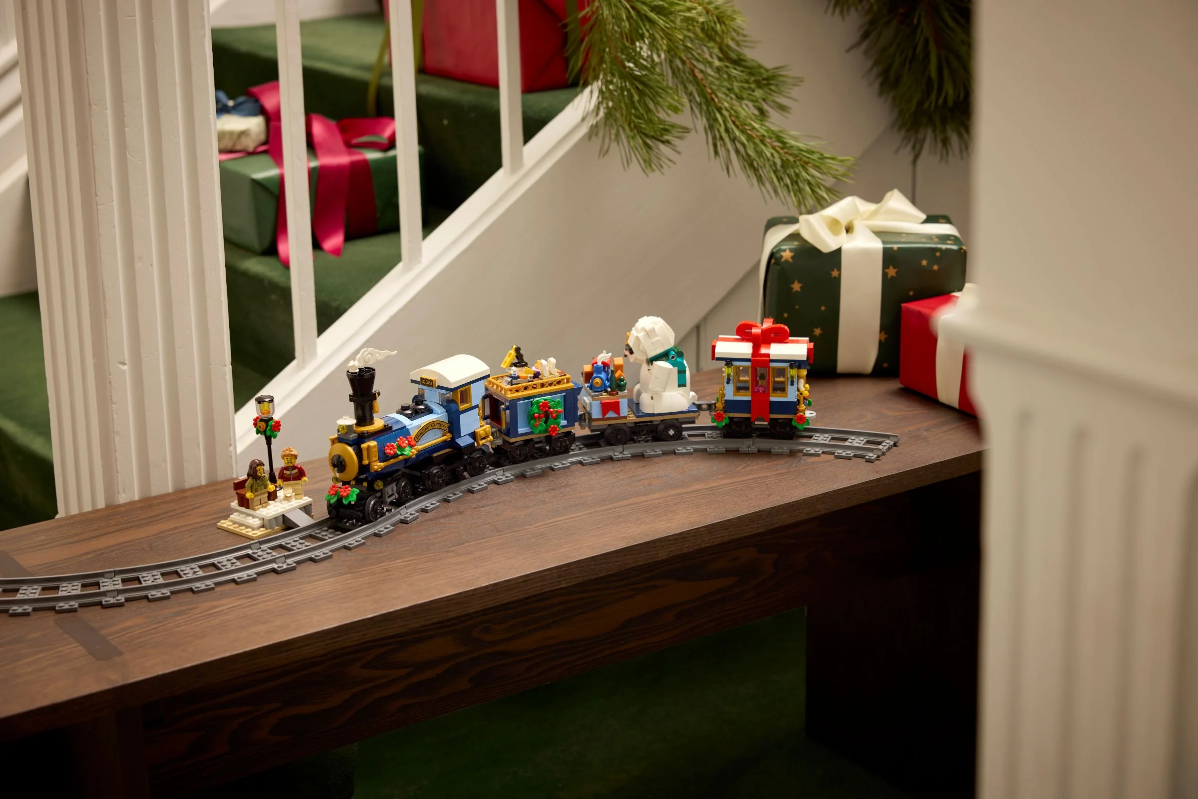 Trenul festiv LEGO 10361 pe șine, lângă cadouri de Crăciun și decorațiuni. Atmosferă magică de sărbători.