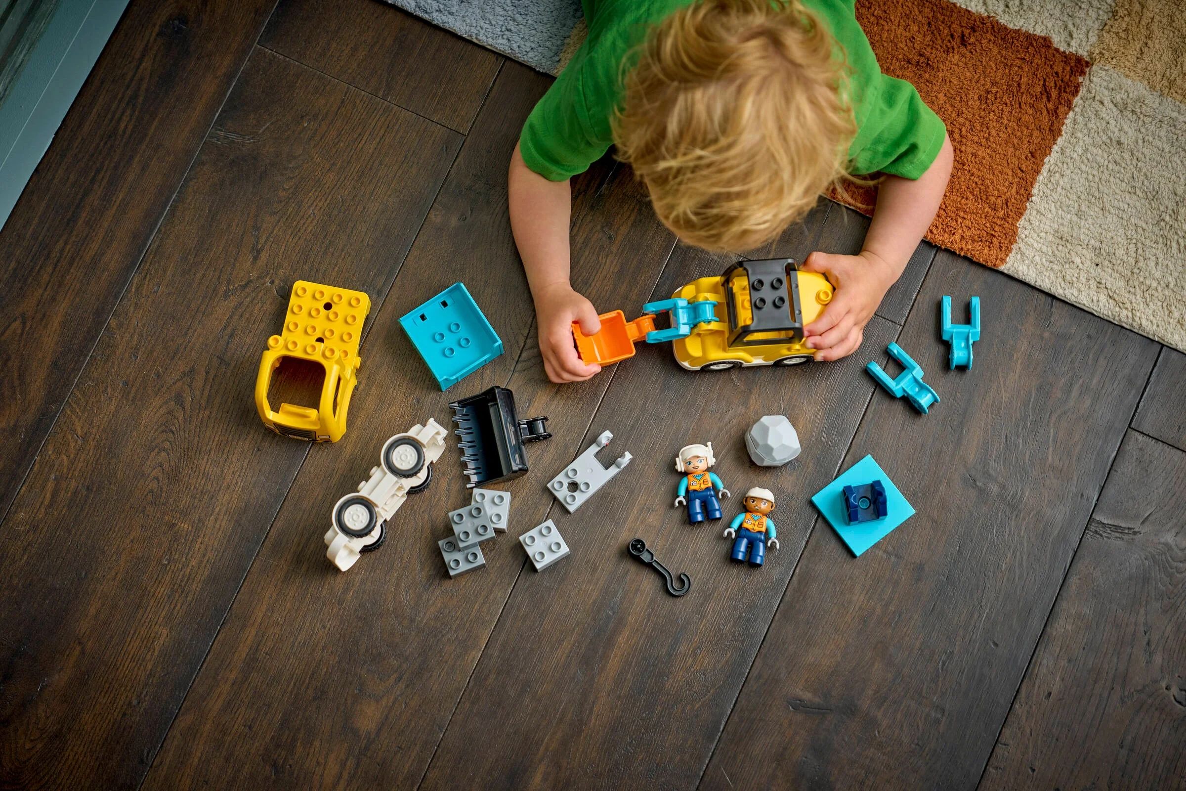 Copil se joacă cu vehiculele de construcție Duplo pe podea. Setul include figurine și diverse componente.