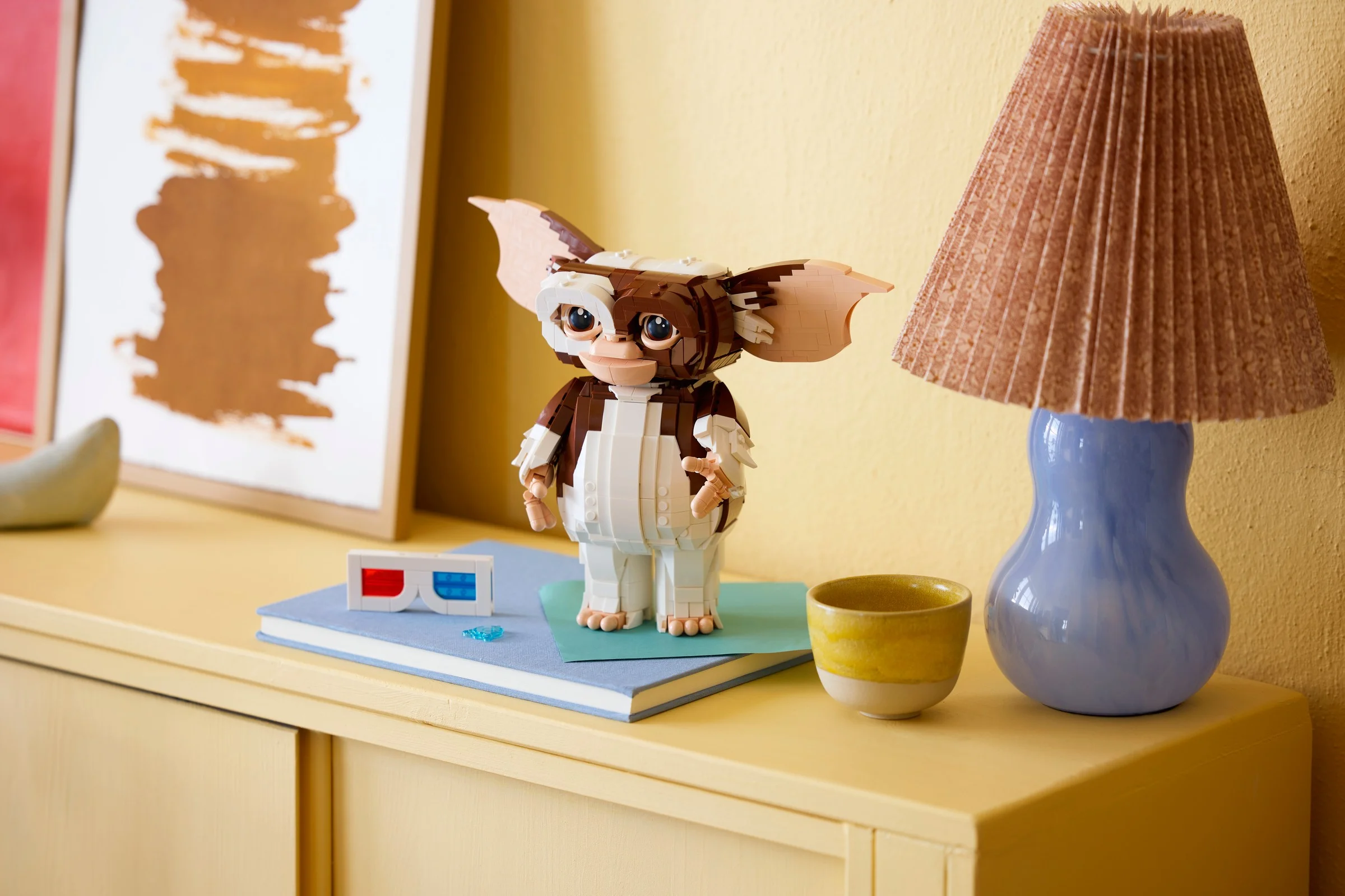 Gizmo LEGO stând pe o carte, alături de ochelari 3D și o lampă. O scenă drăguță!