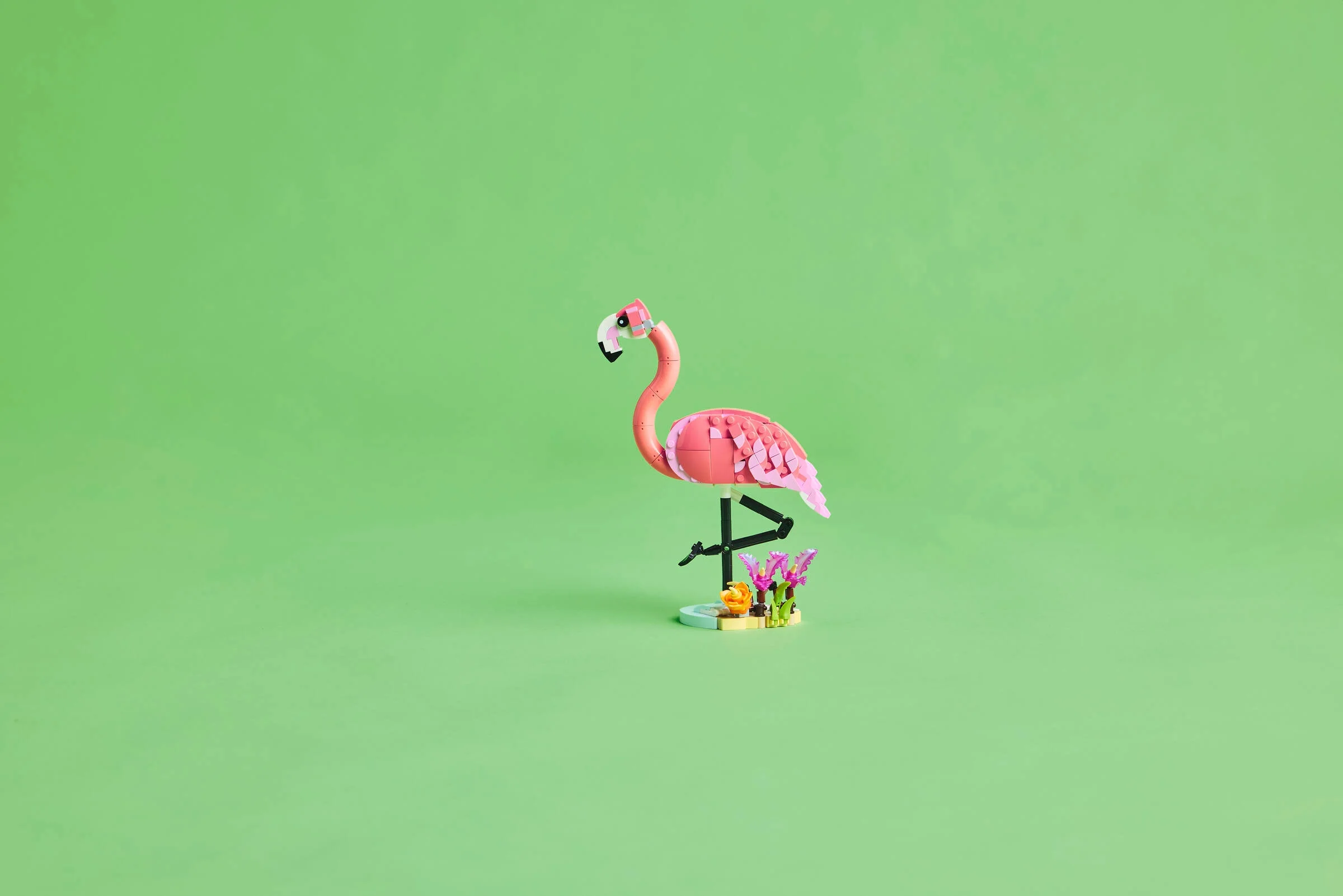 Un flamingo roz LEGO stând pe un picior, cu flori și o bază albastră-verzuie.
