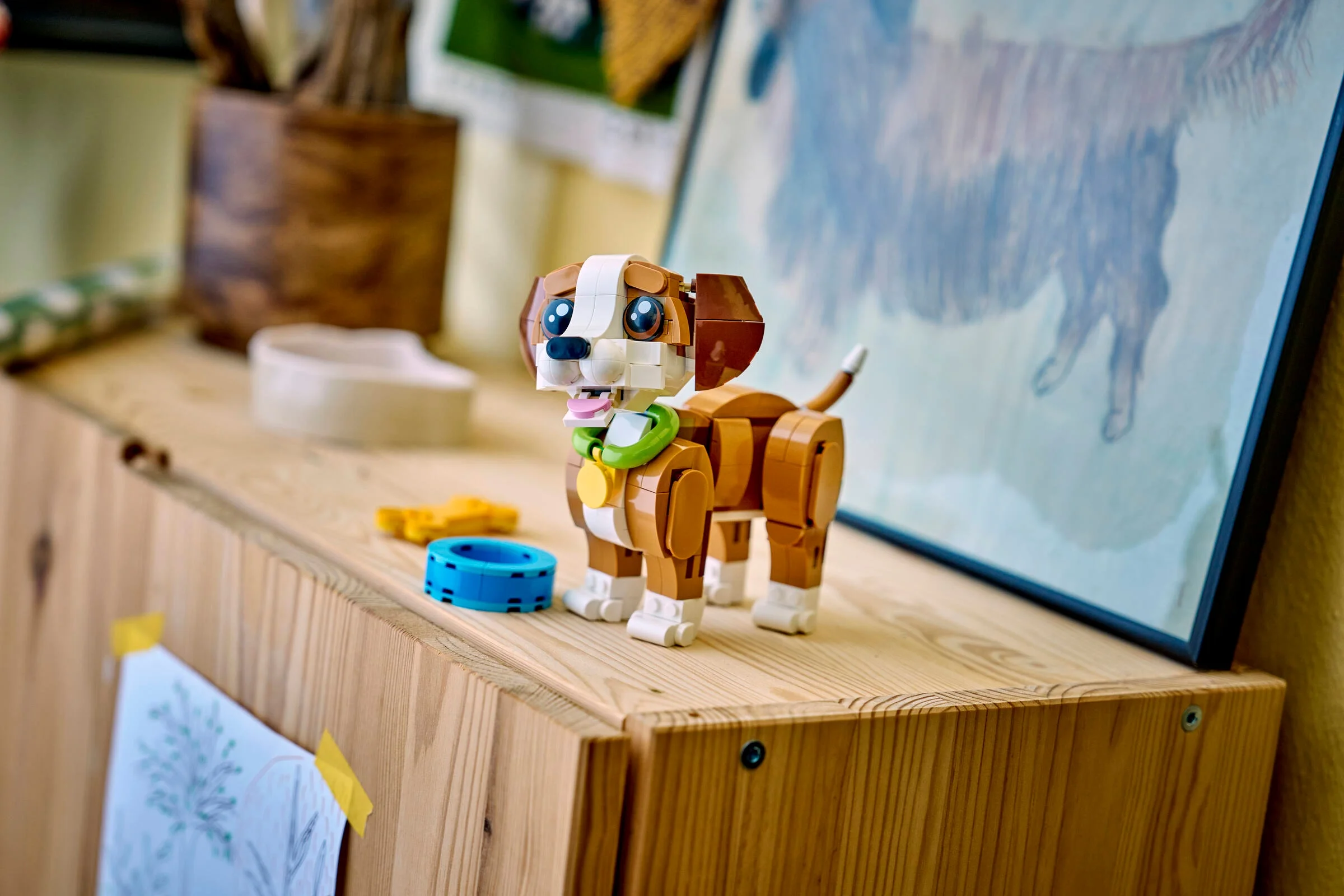 Cățelușul LEGO maro și alb stă jucăuș pe o masă de lemn. Jucării și un tablou în fundal.