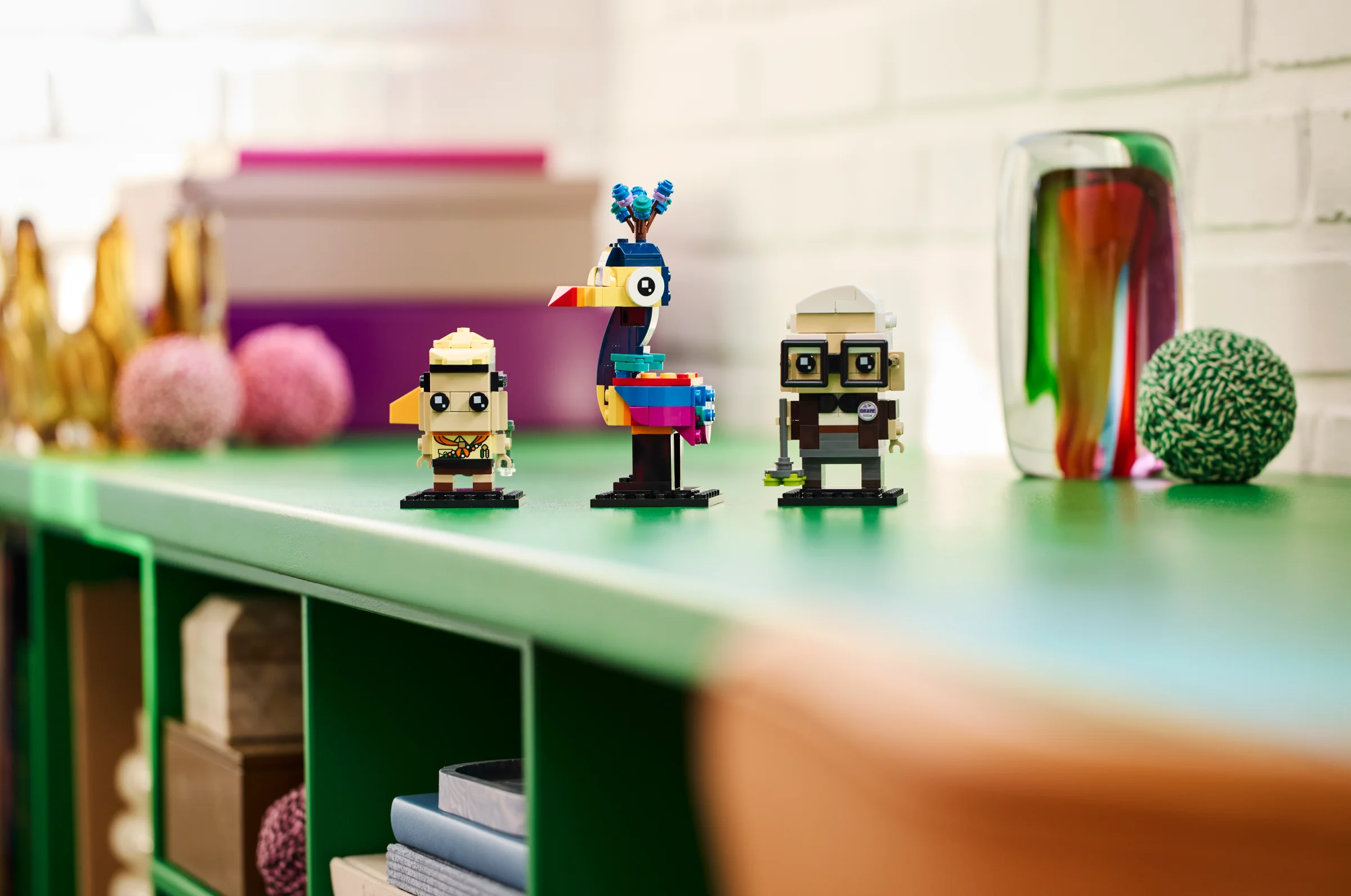Prezentare cu figurinele BrickHeadz Carl, Russell și Kevin din filmul Up. Sunt așezate pe un raft verde.