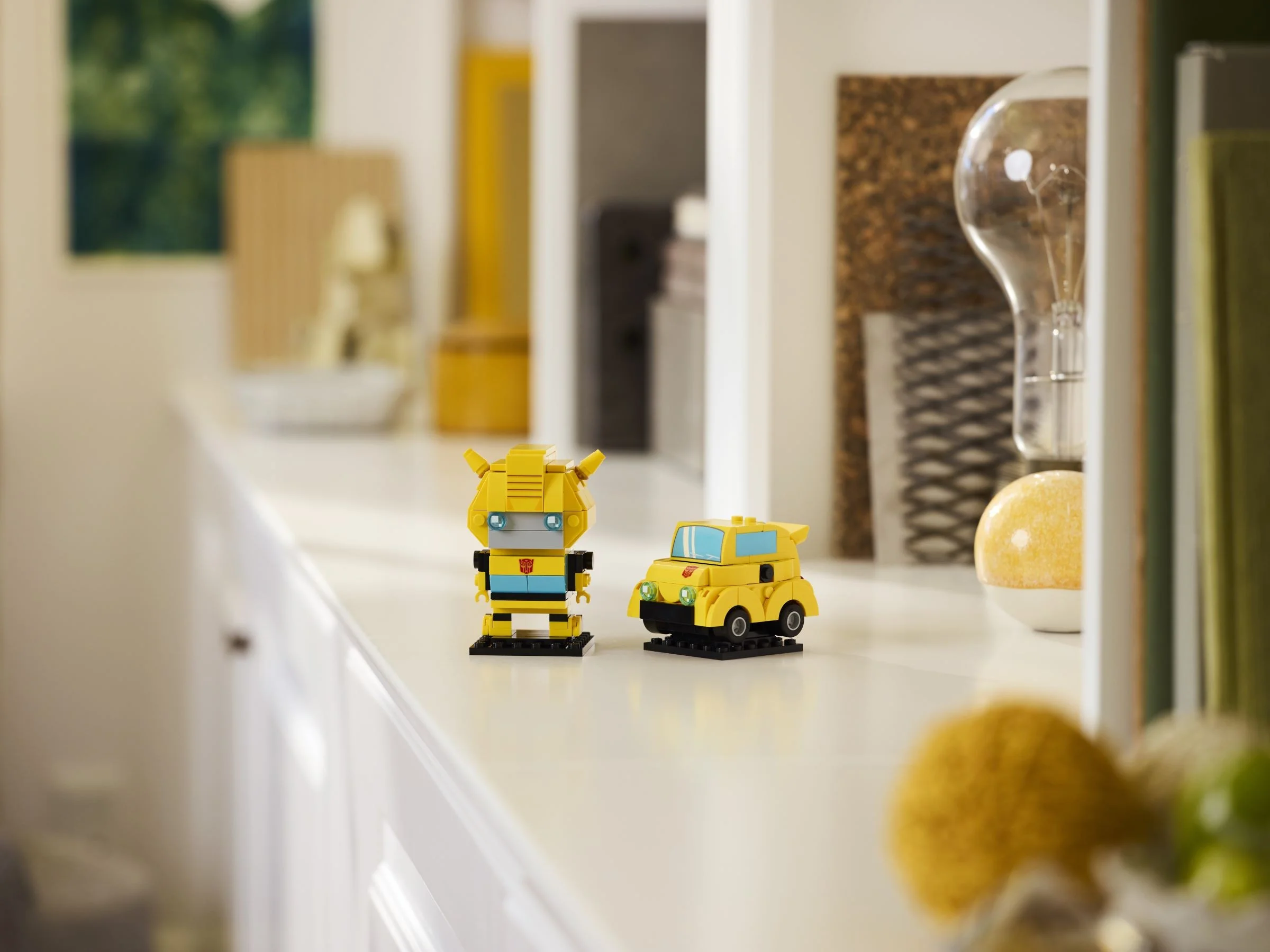 Imagine cu Bumblebee BrickHeadz: robot și mașină galbenă. Construcție LEGO detaliată, pe o suprafață albă.