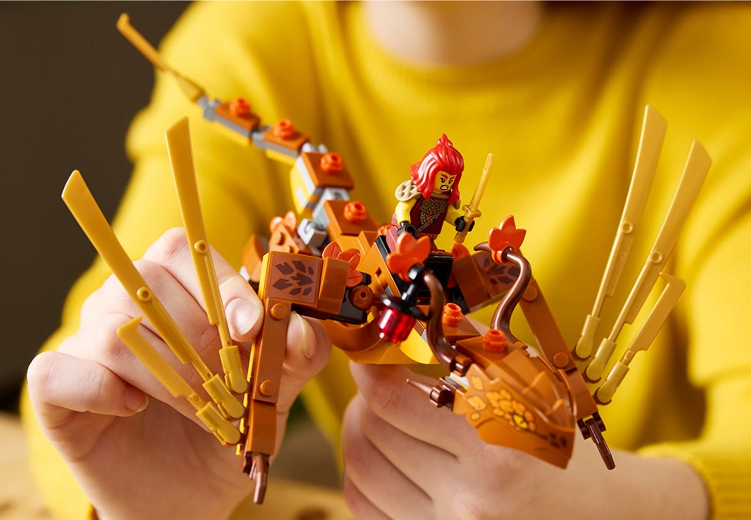 Imagine cu o minifigurină LEGO Ninjago pe un dragon portocaliu, ținut în mâini.