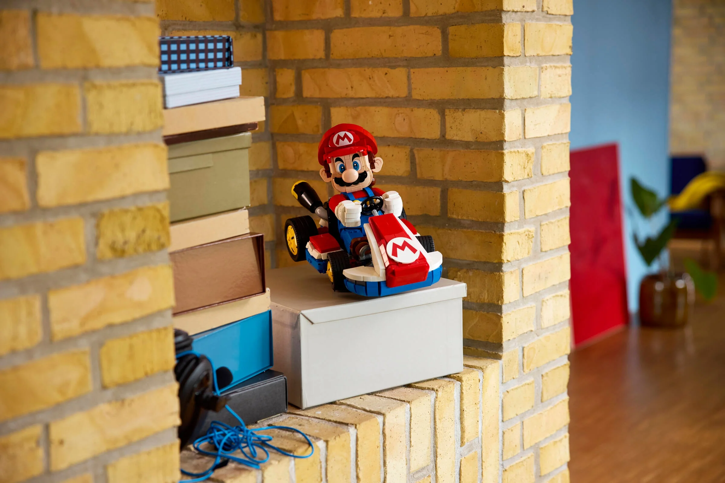 Mario în kartul său LEGO, așezat pe o cutie albă, lângă un perete de cărămidă galbenă.