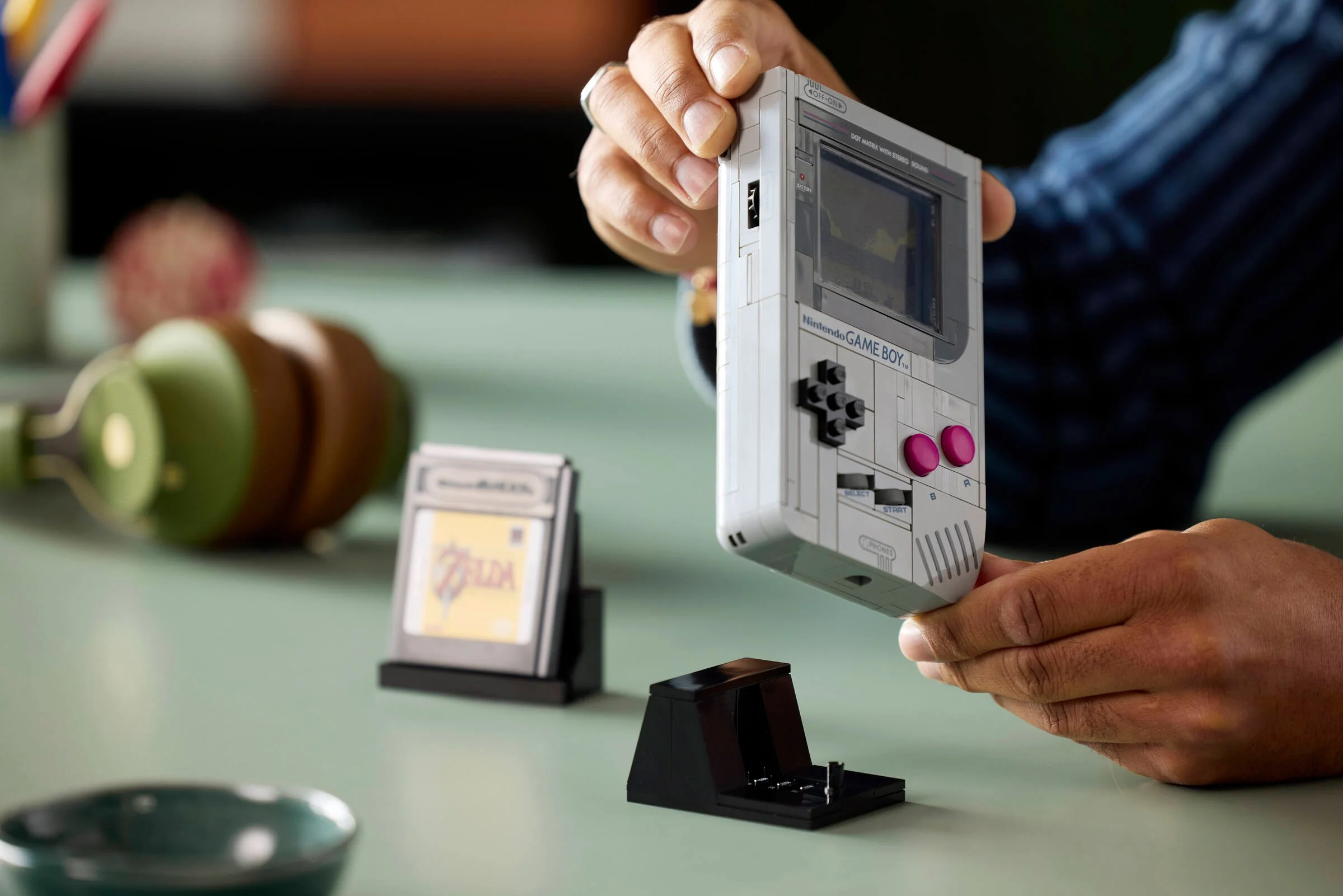 Imagine cu LEGO Game Boy ținut în mână, cartuș Zelda și suport. Detalii.