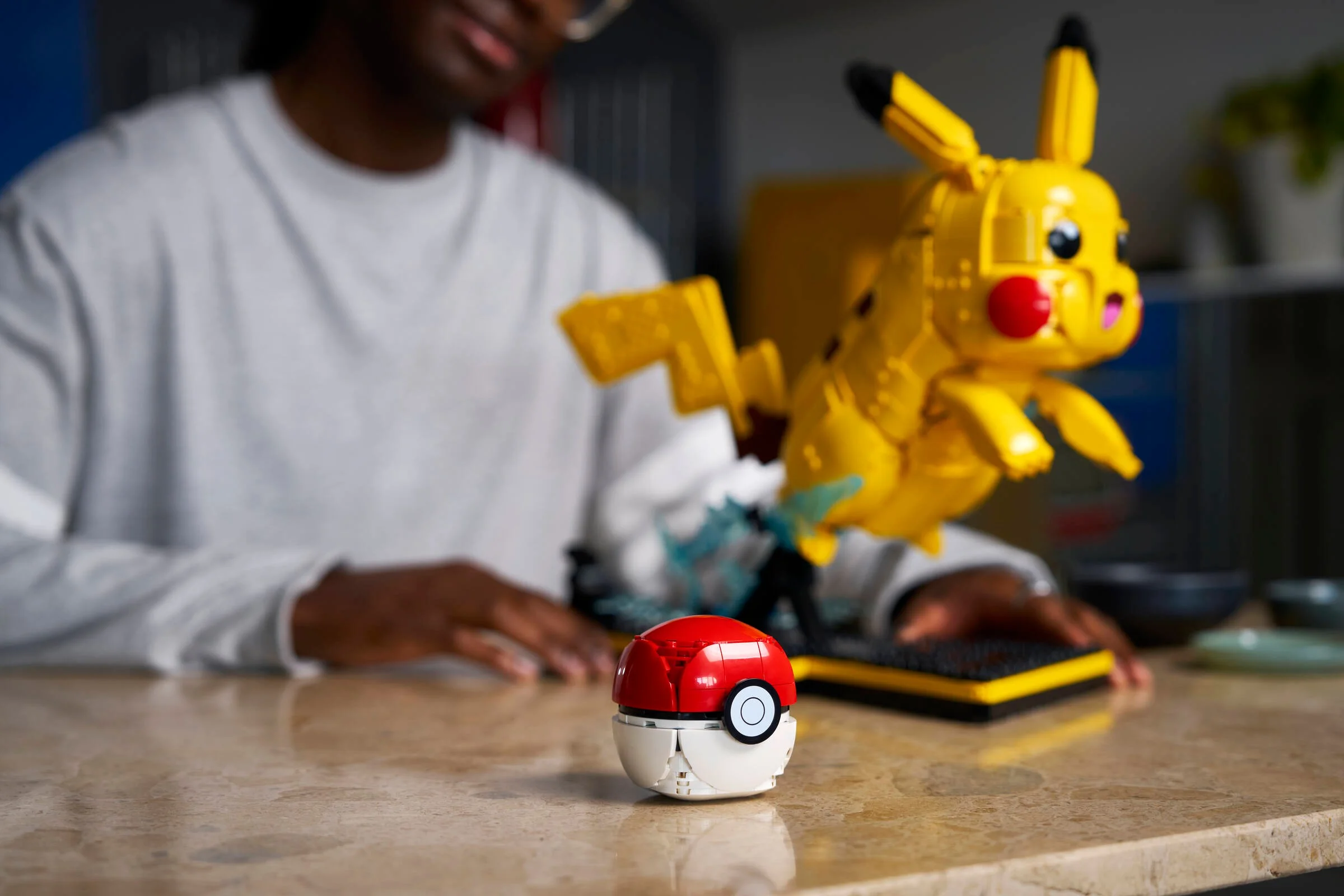 Imagine cu Pikachu și Poké Ball construite din LEGO, pozate pe o masă, cu o persoană în fundal.