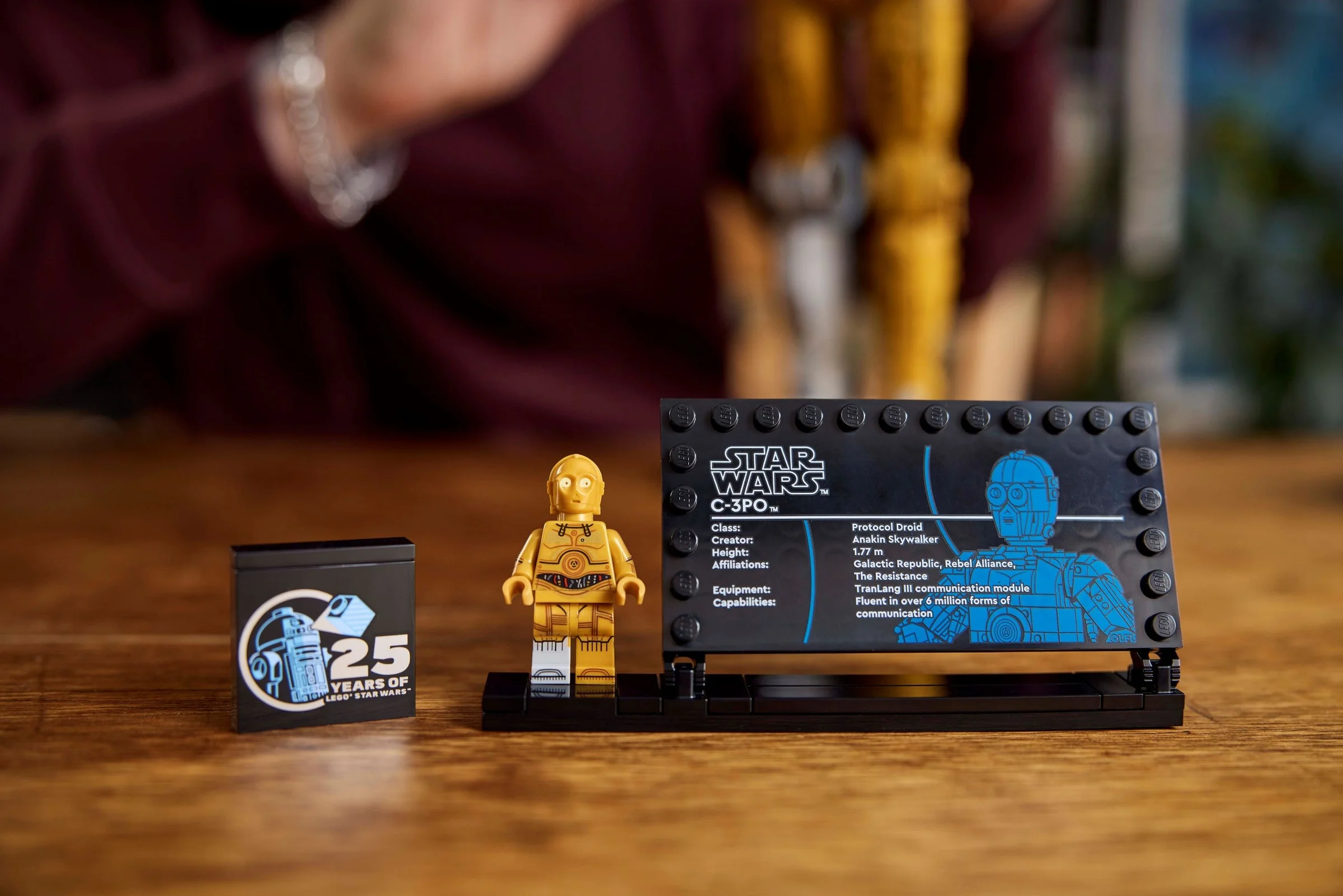 Imagine cu minifigurina C-3PO, placa informativă și cărămida aniversară, pe un fundal cald de lemn.