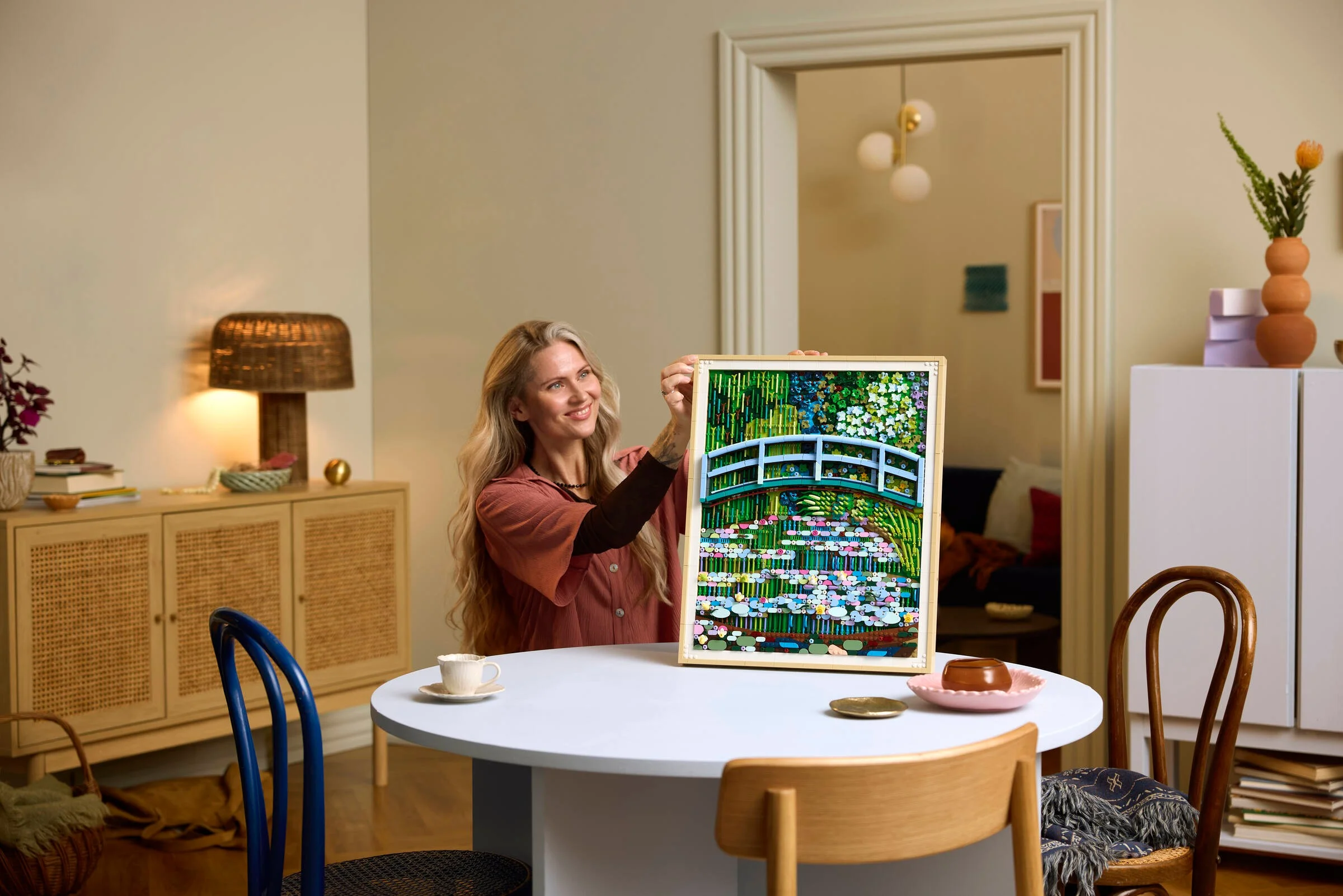 Femeie zâmbitoare ținând tabloul LEGO Monet, adăugând o notă artistică interioarelor moderne.