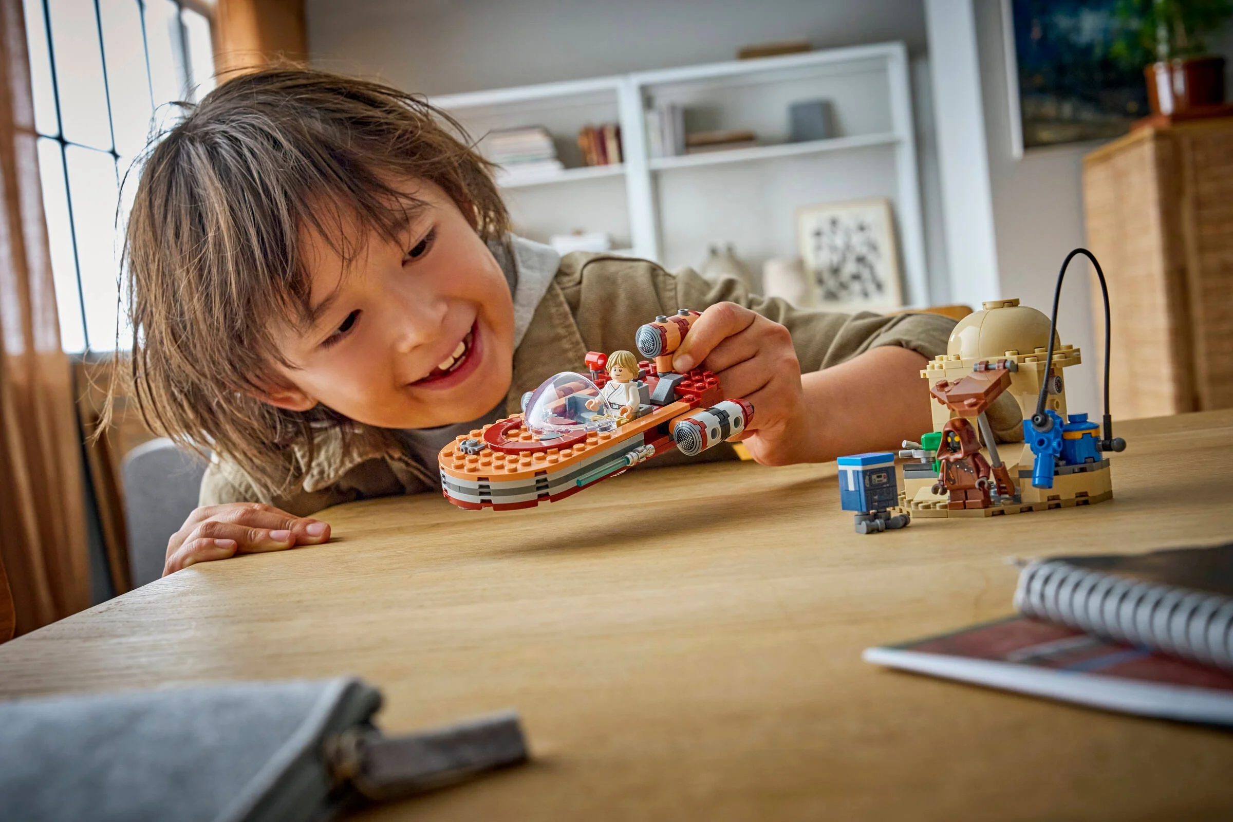Un copil zâmbește jucându-se cu setul LEGO Landspeeder, incluzând minifigurine și detalii ale modelului.