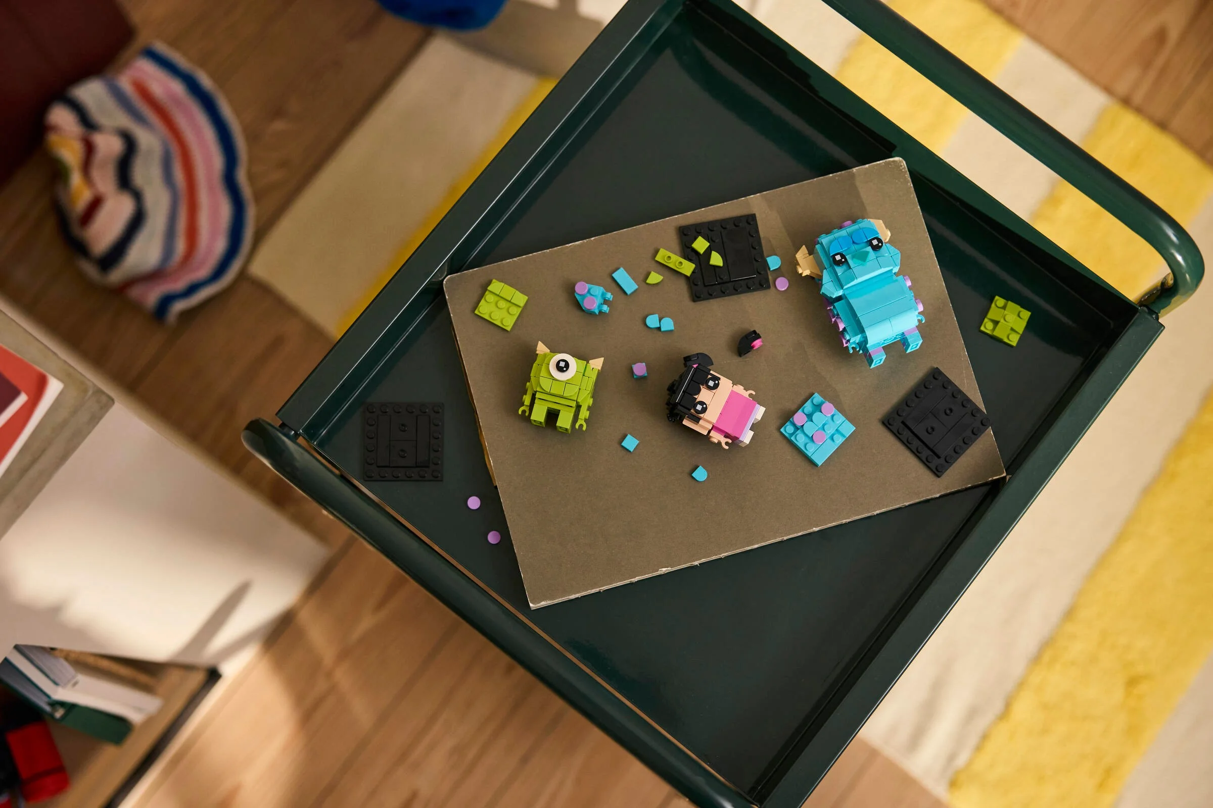 O imagine de sus cu figurile Sulley, Mike și Boo BrickHeadz LEGO, parțial construite pe o măsuță verde.