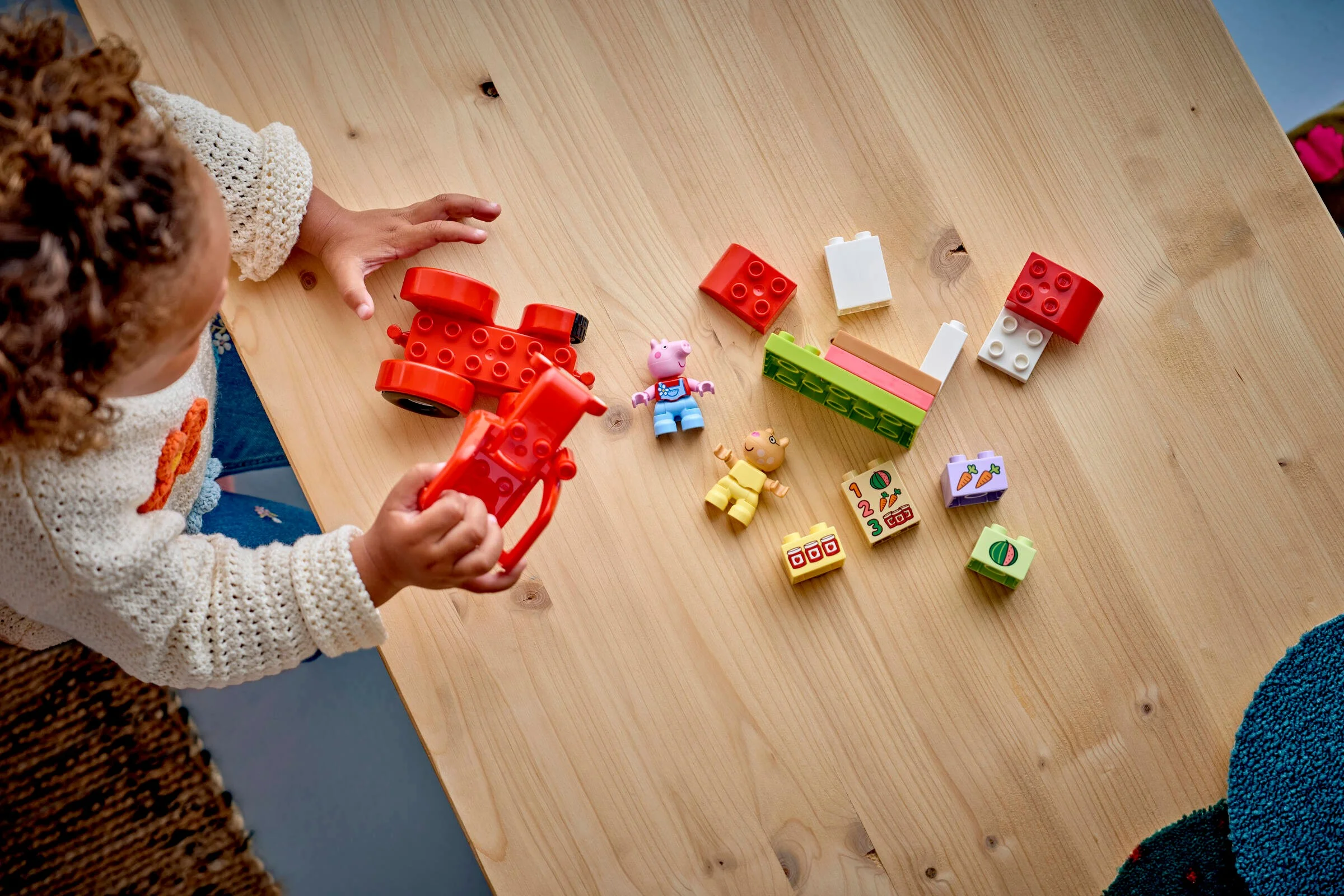 Copil joacă cu tractorul și setul de piață Duplo, creând o scenă distractivă. Include figurine și cărămizi colorate.