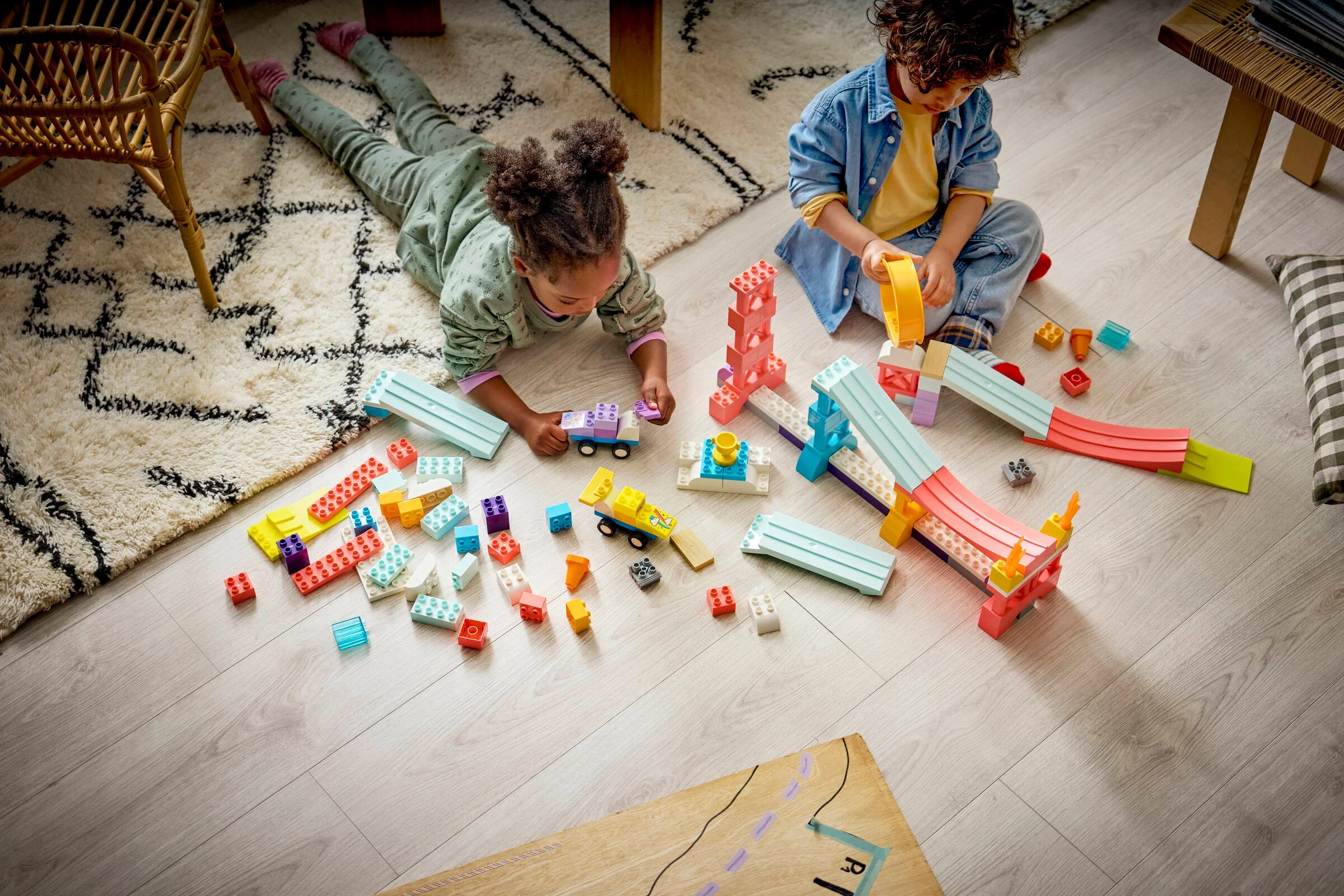 Doi copii se joacă cu rampe și vehicule Duplo, construind pe podea. Joacă creativă și colorată.