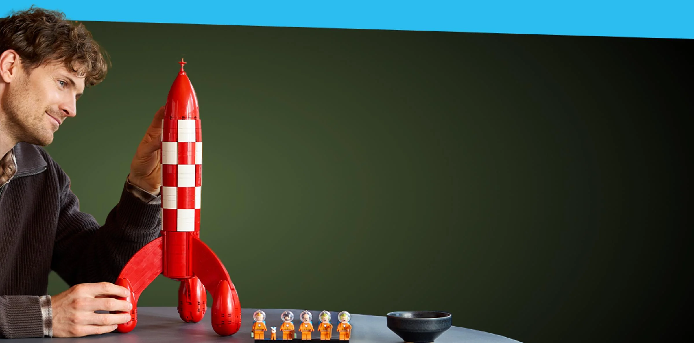 Bărbat admirând racheta Tintin LEGO, cu minifigurine și o ceașcă pe masă.