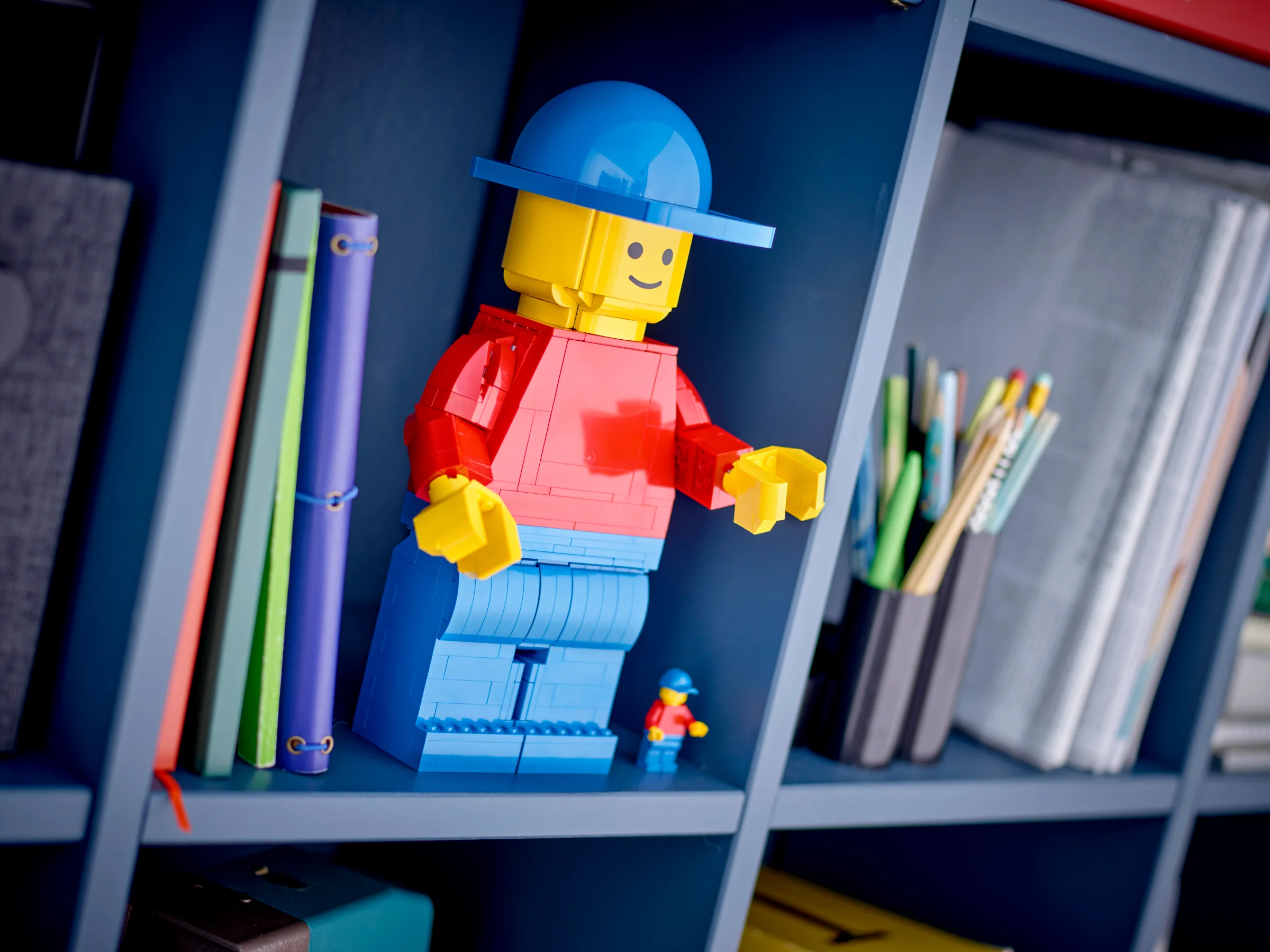 Figurină LEGO mărită, cu căciulă albastră, pe un raft. Alături, o minifigurină standard, ca referință.