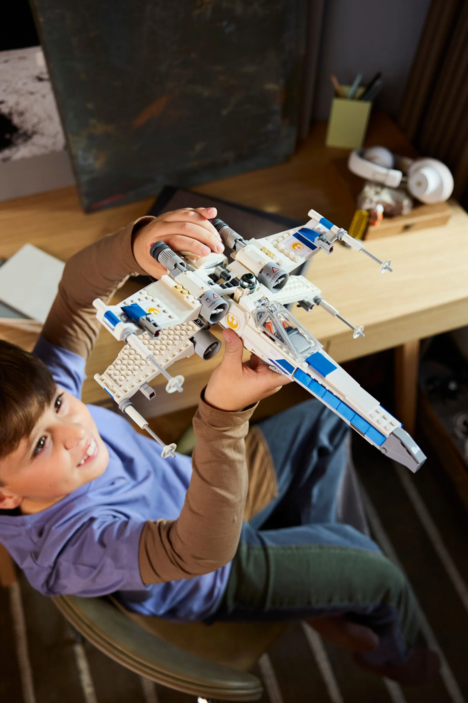 Un băiat ține în mâini X-wing-ul LEGO, zâmbind. Modelul este detaliat și colorat.