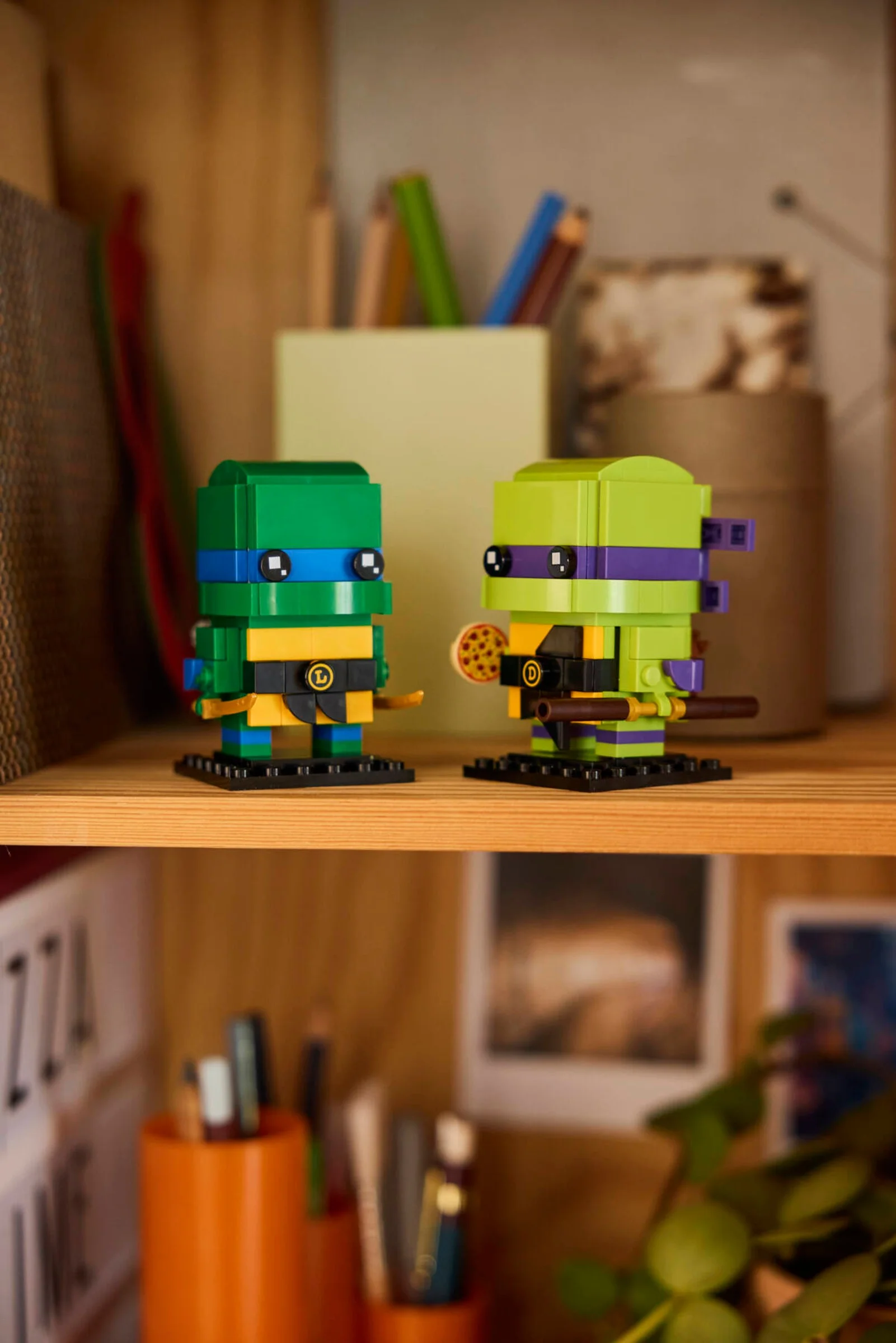 Două figurine BrickHeadz TMNT, Leonardo și Donatello, pe un raft. Fundal cu obiecte diverse.