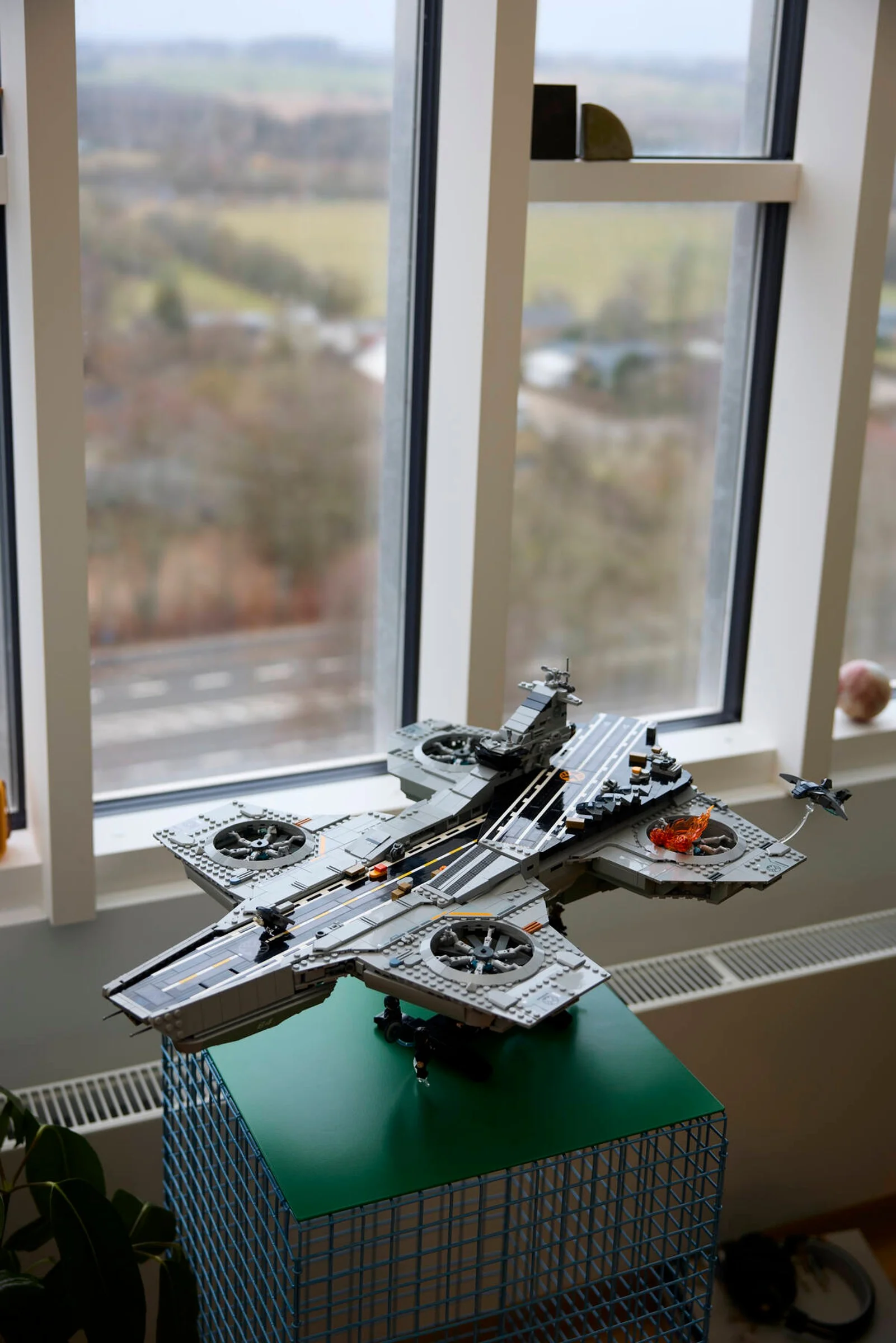 Imagine cu Helicarrier-ul S.H.I.E.L.D. LEGO, plasat lângă o fereastră. Model complex, detaliat.