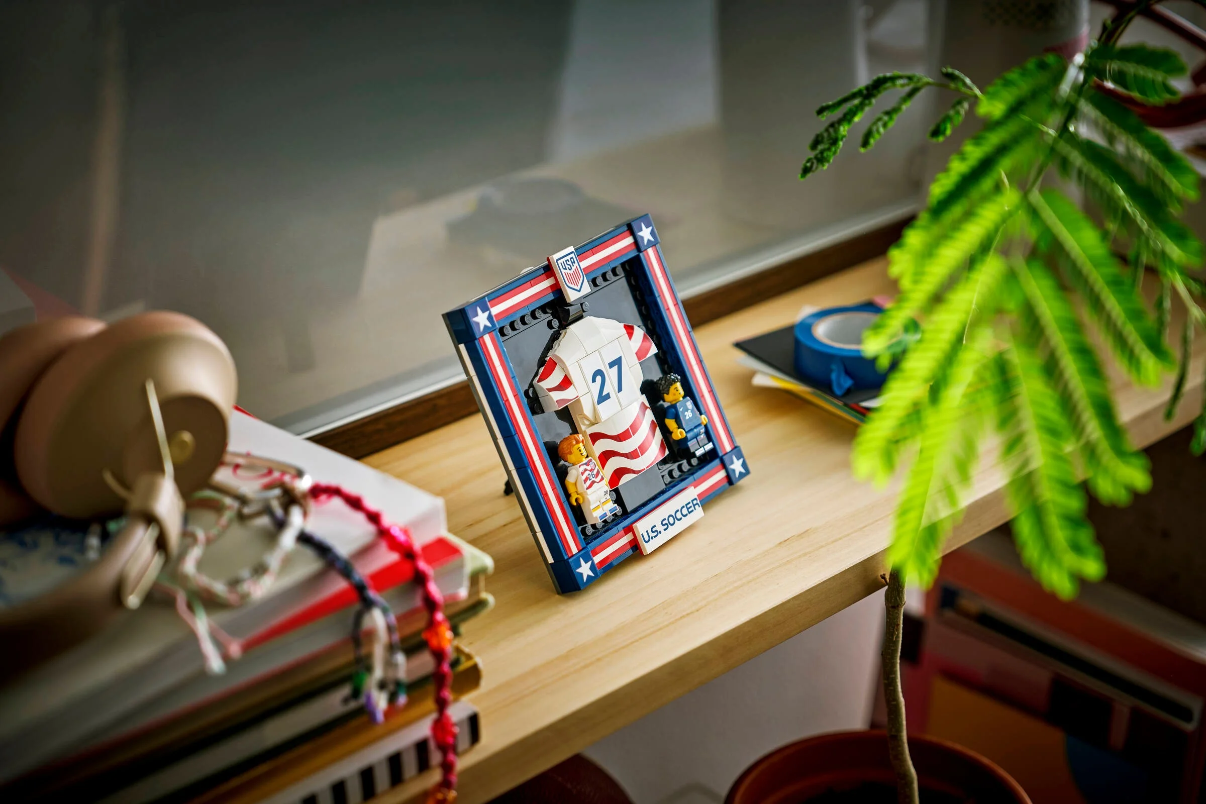 Setul LEGO U.S. Soccer Jersey pe un raft, lângă căști și o plantă.