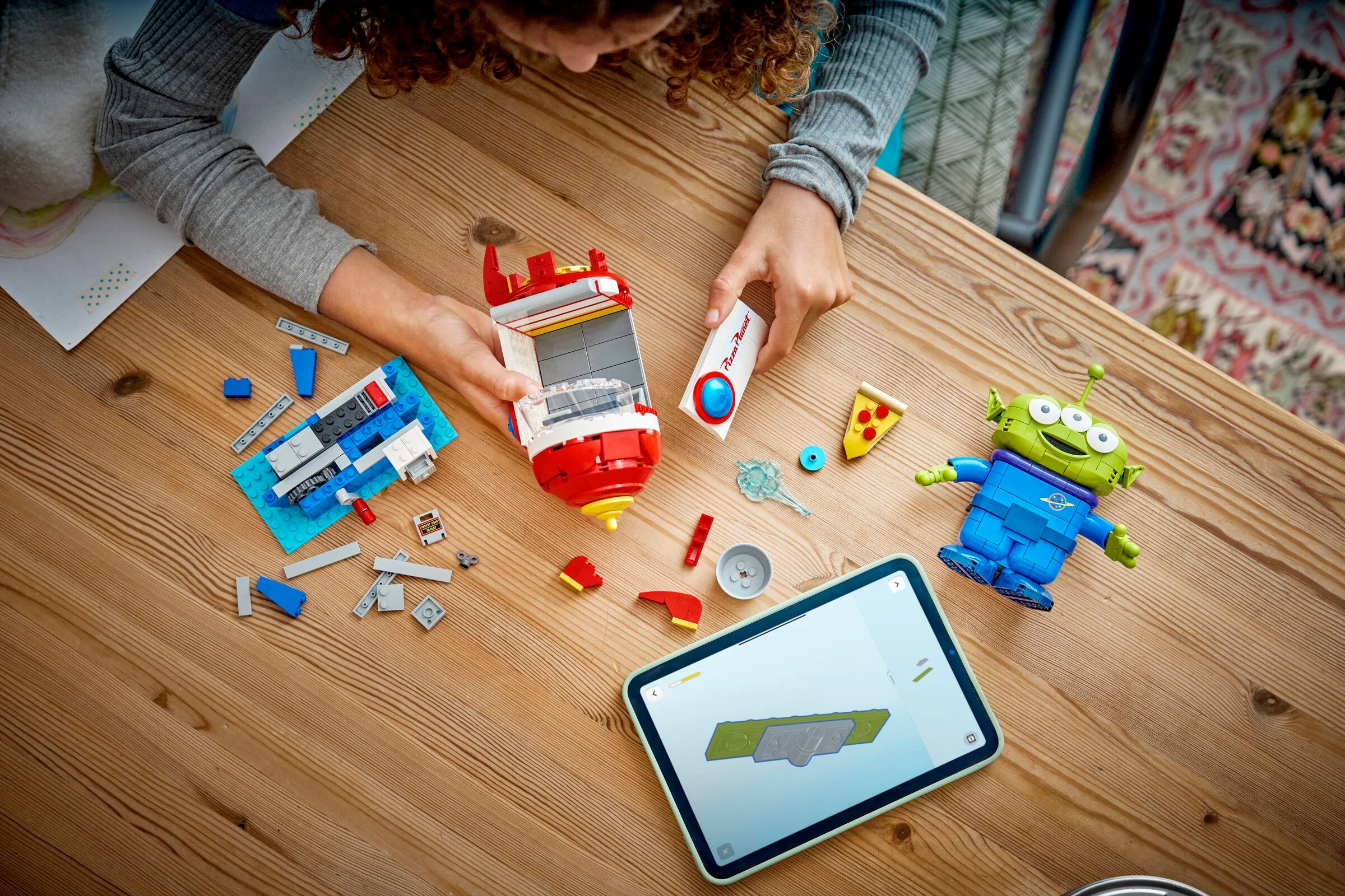 Copil asamblând setul LEGO Alien cu rachetă Pizza Planet. Piesele și instrucțiunile digitale sunt vizibile.