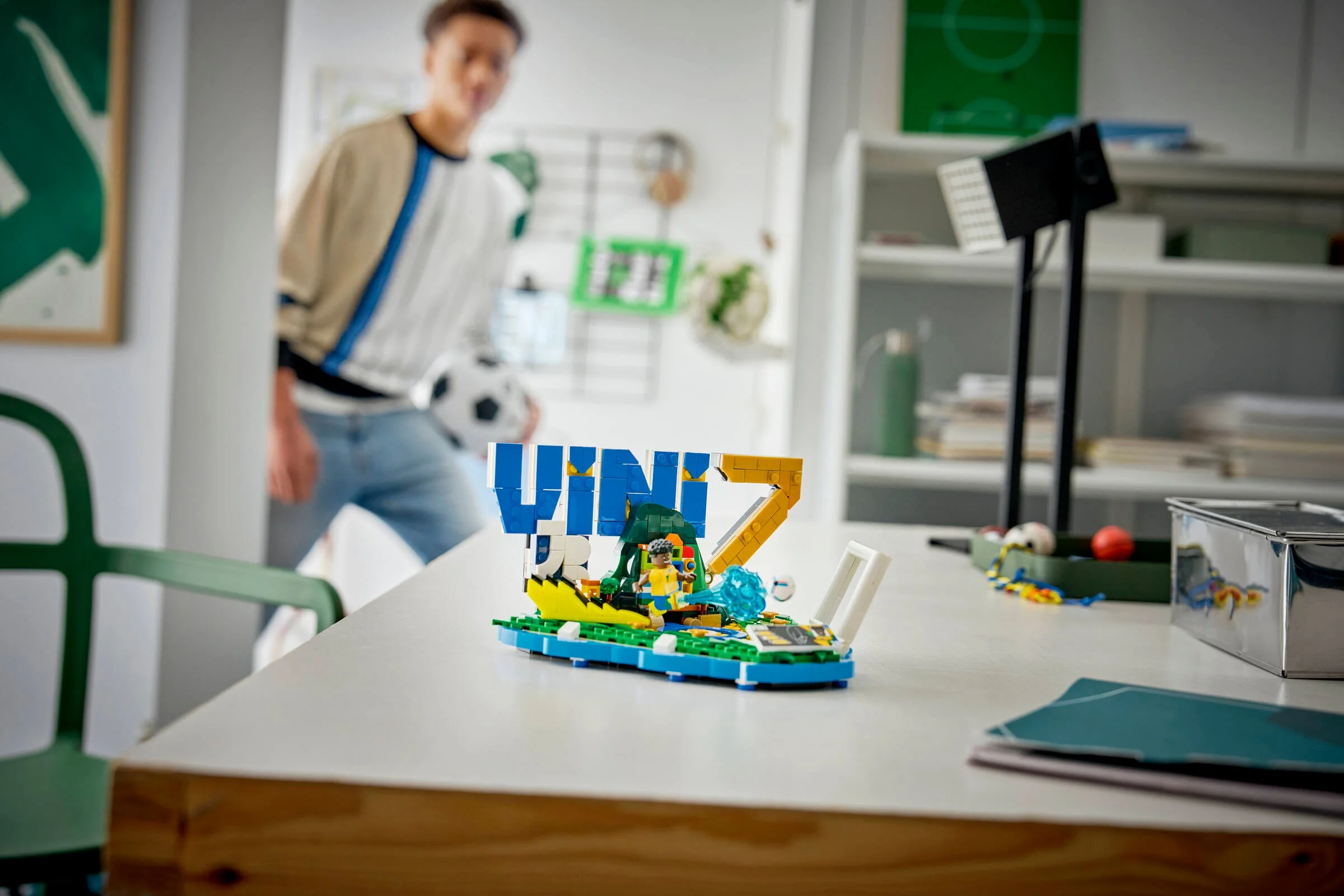 Prezentare a setului LEGO Vini Jr. – Football Highlights pe un birou, cu un tânăr în fundal.