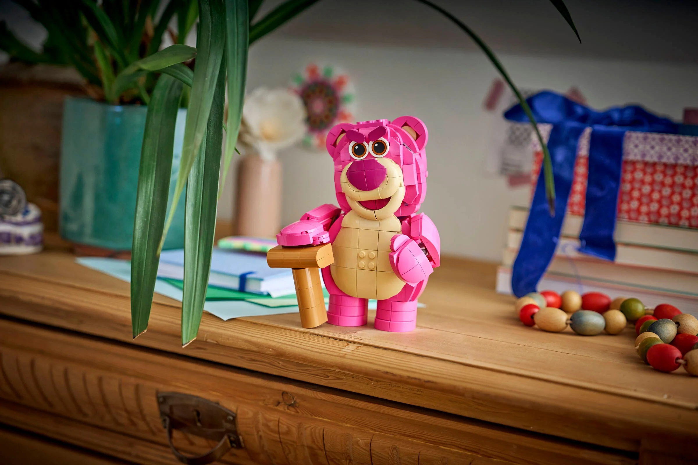 Lotso, ursulețul roz construit din LEGO, stând pe o comodă din lemn, decorat cu plante.