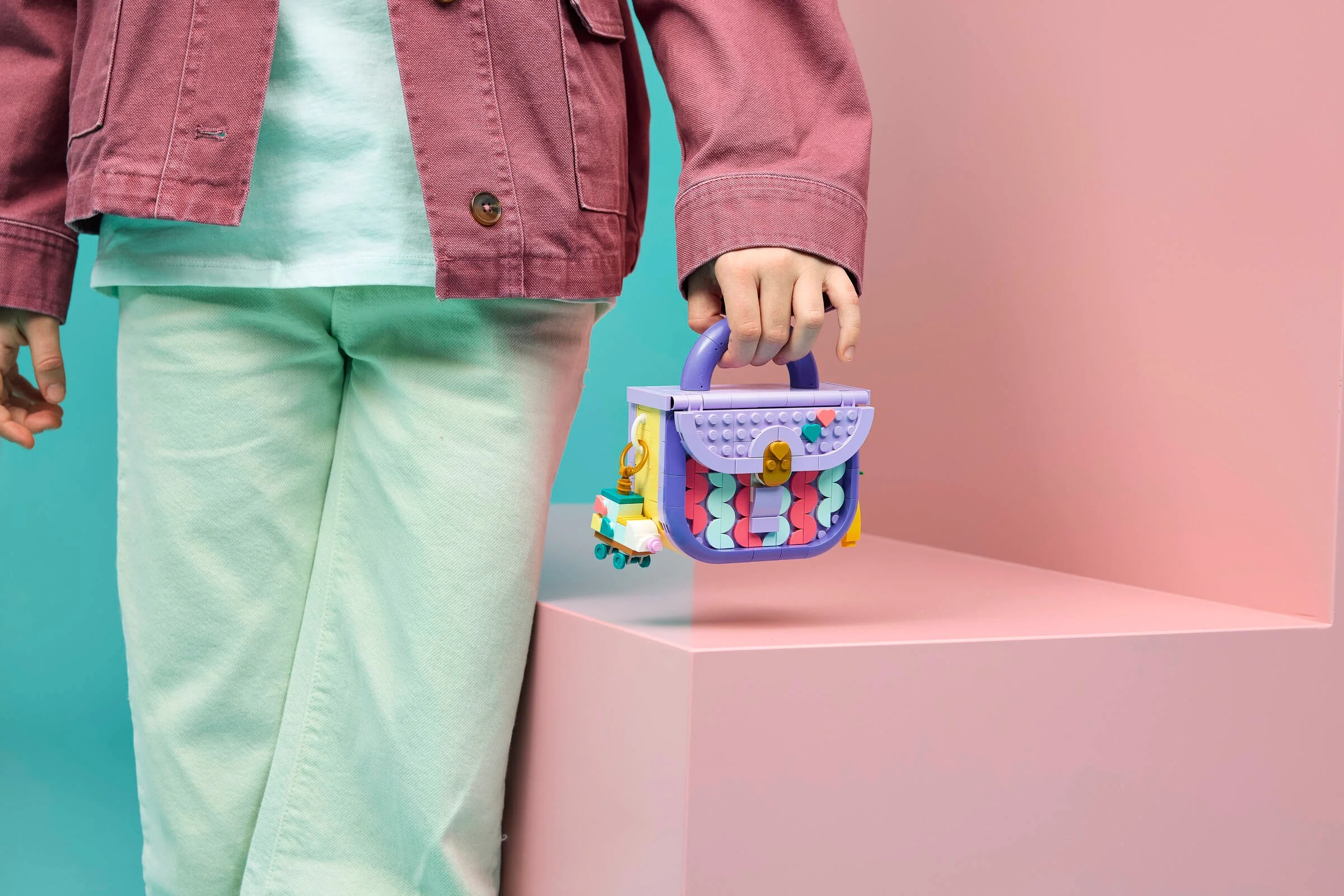 Geantă LEGO viu colorată ținută de o persoană, pe un fundal roz și albastru.