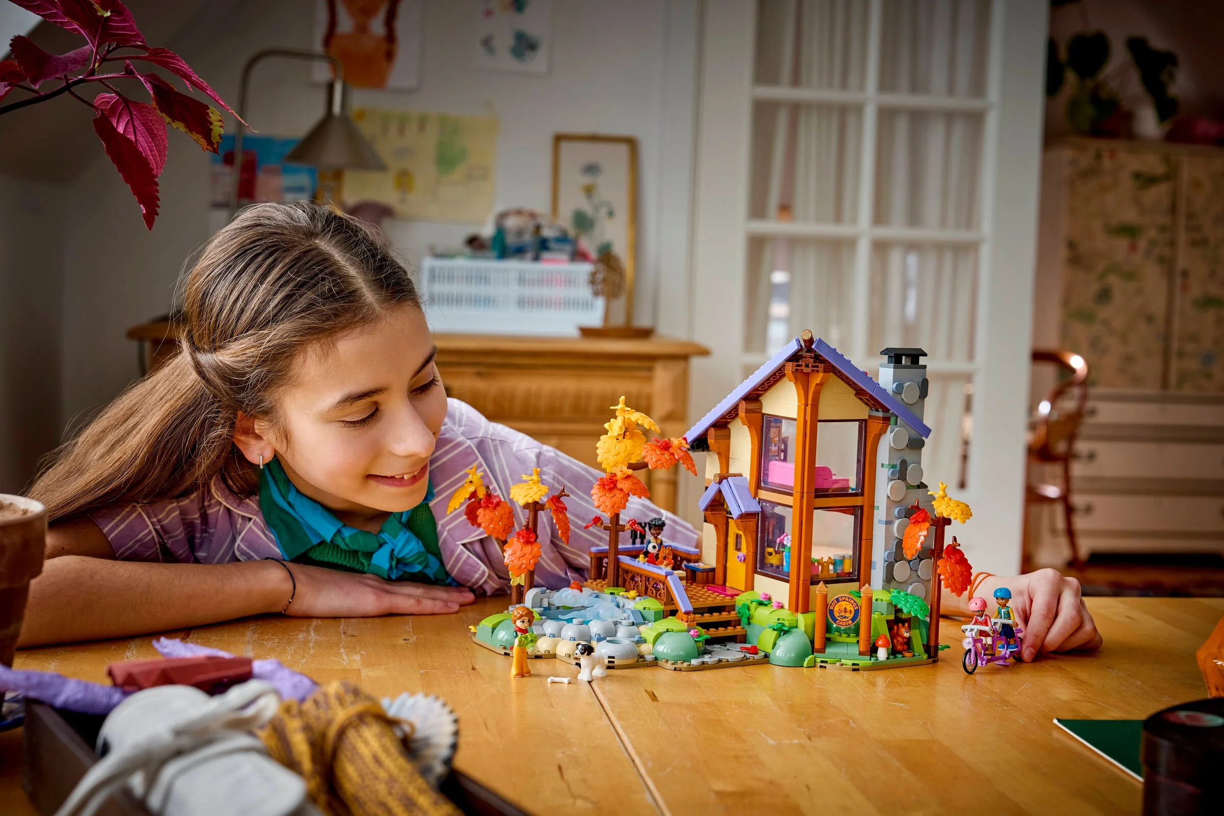O fată zâmbește admirând cabana LEGO, într-o atmosferă autumnală. Model detaliat.