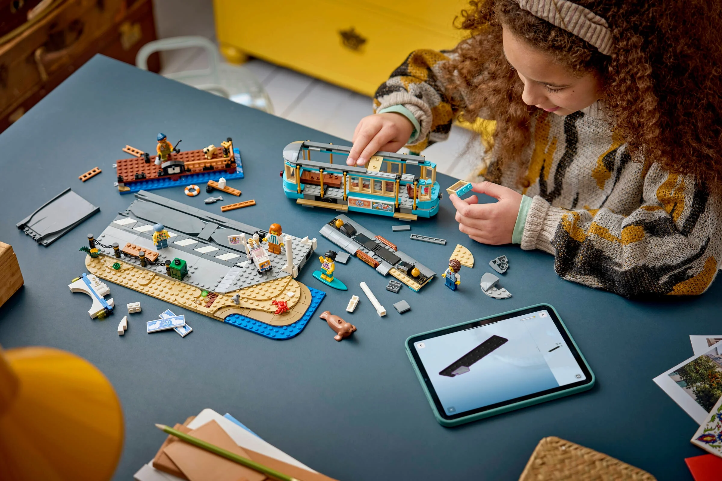 Fată construind tramvaiul de plajă LEGO Classic. Model parțial asamblat și instrucțiuni digitale pe tabletă.