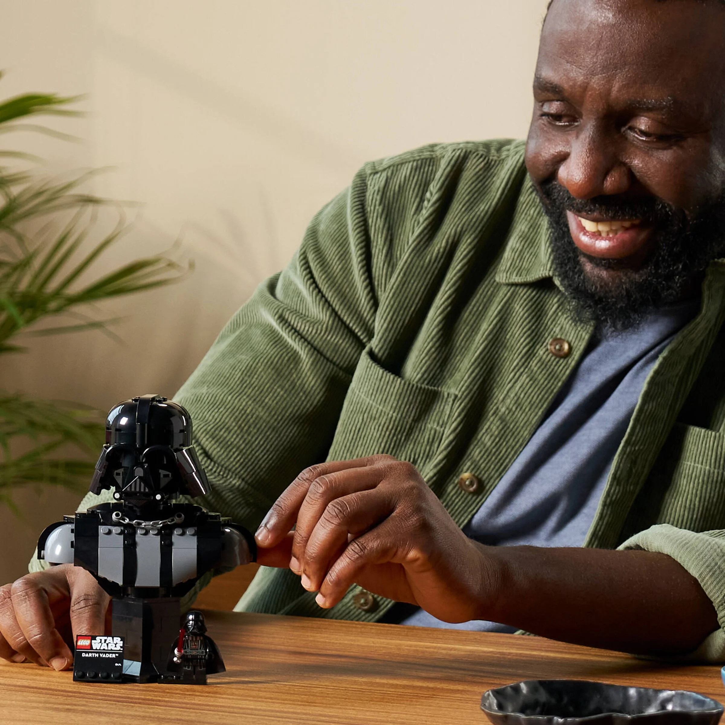 Un bărbat zâmbește admirând bustul LEGO Darth Vader construit, pe o masă de lemn.