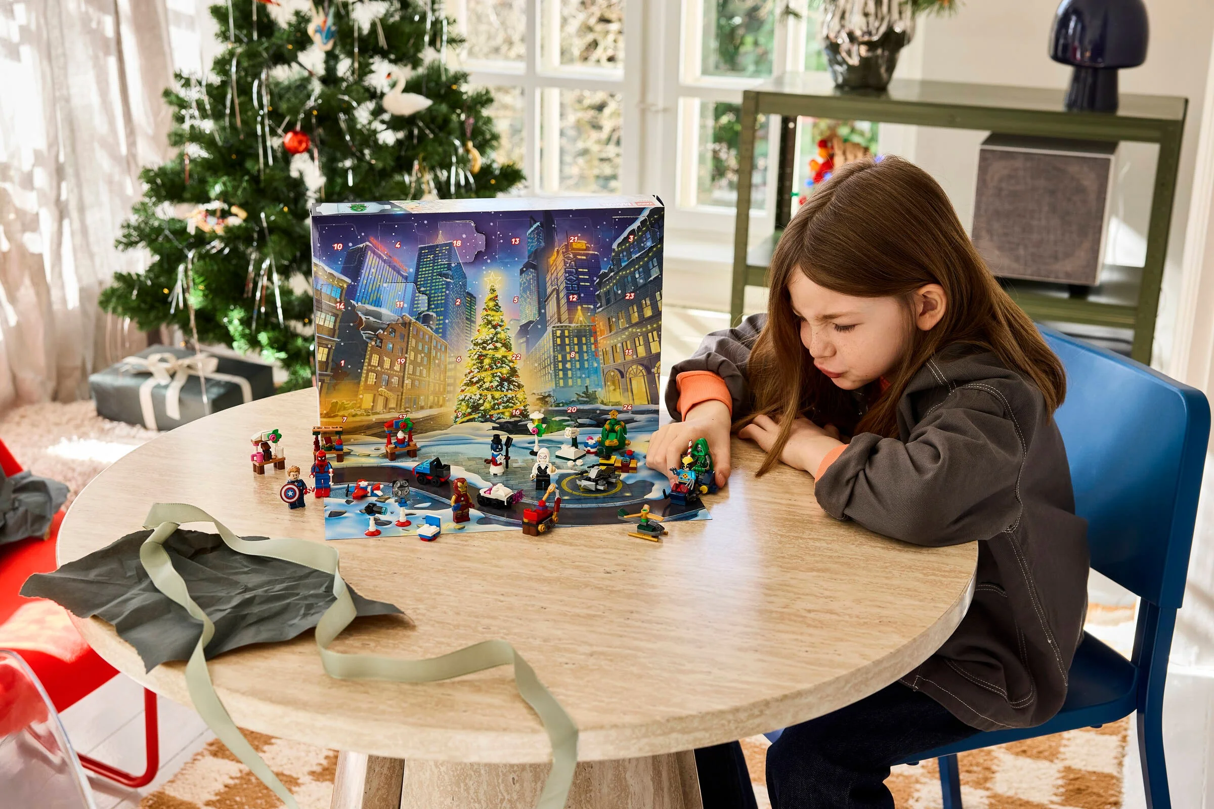 O fetiță se joacă cu setul LEGO Marvel Calendar Advent 2026, lângă un brad împodobit.