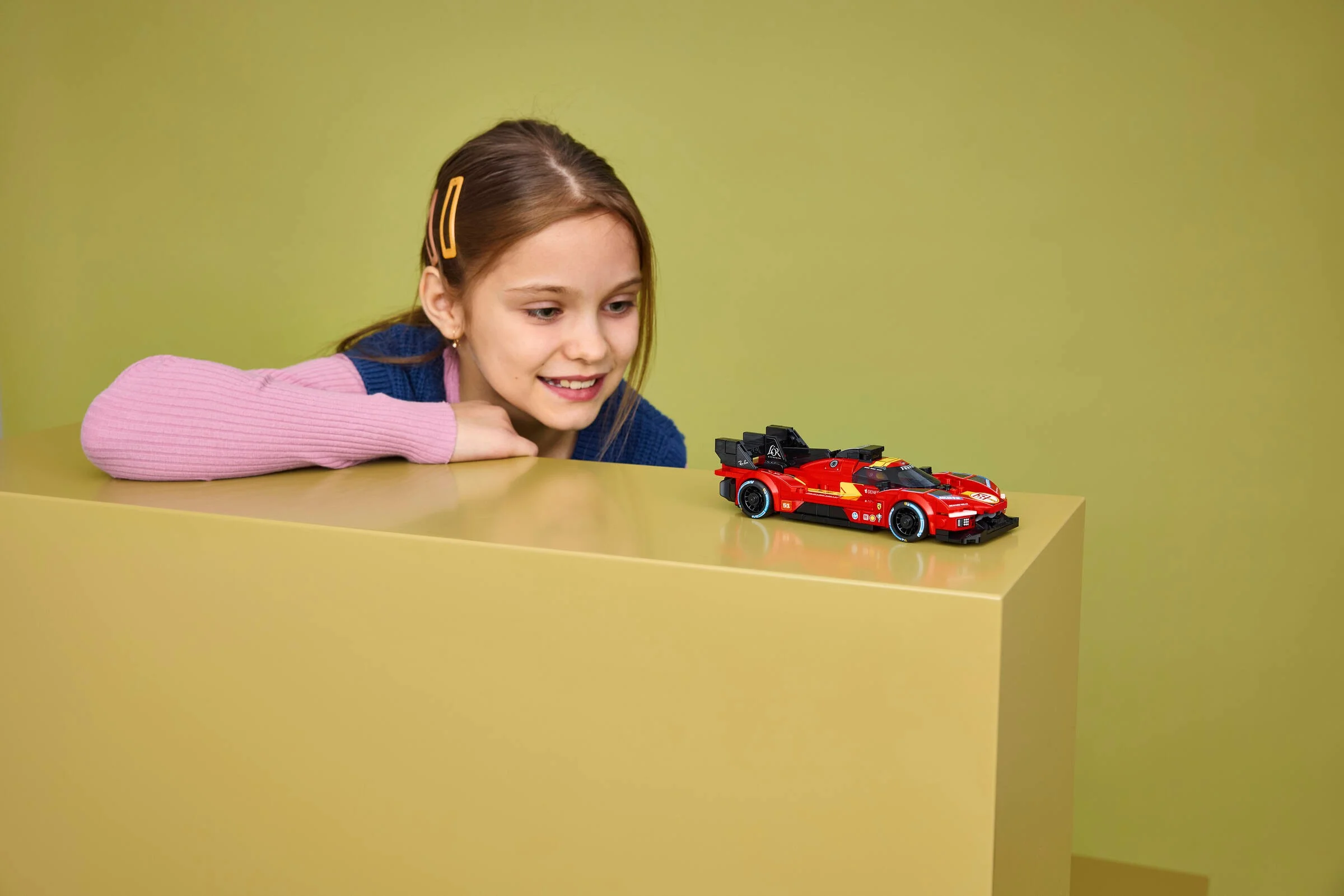O fetiță zâmbește admirativ la modelul Ferrari 499P construit din LEGO. Jucărie atractivă!