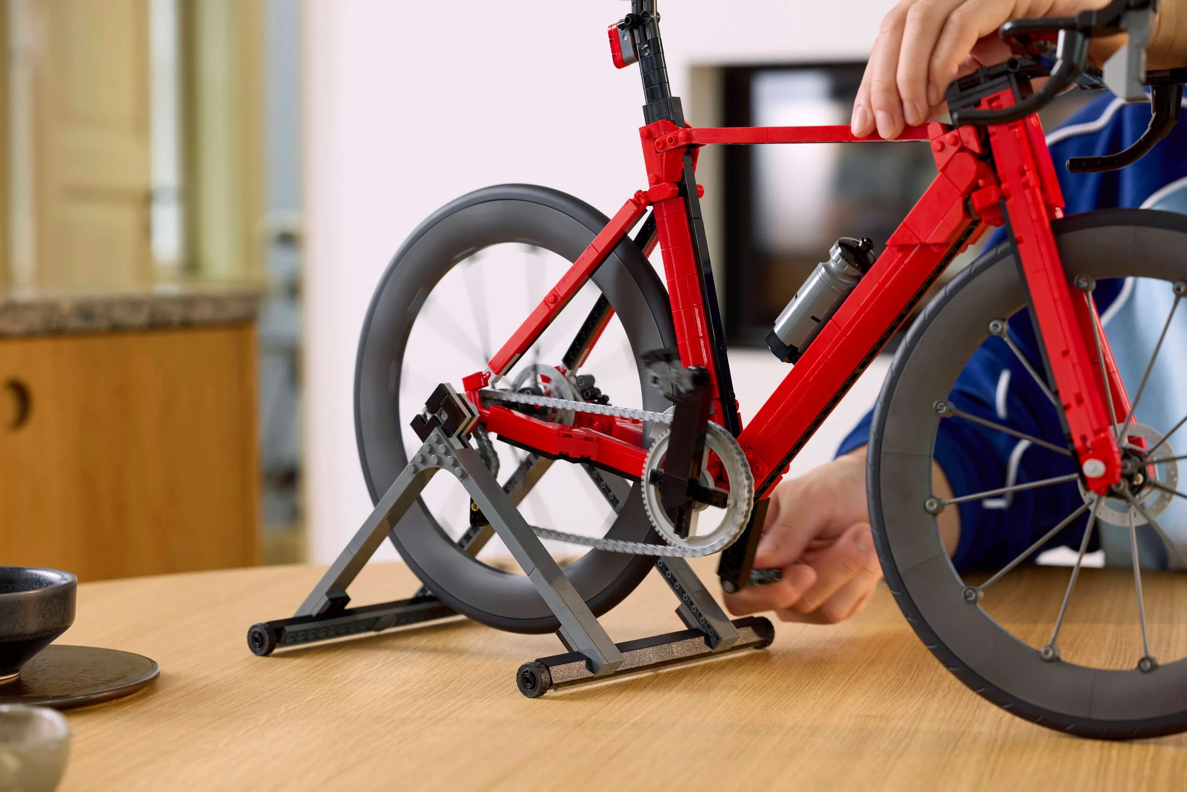 Prim-plan cu bicicleta roșie LEGO pe suport, cu mâini ajustând pedala.