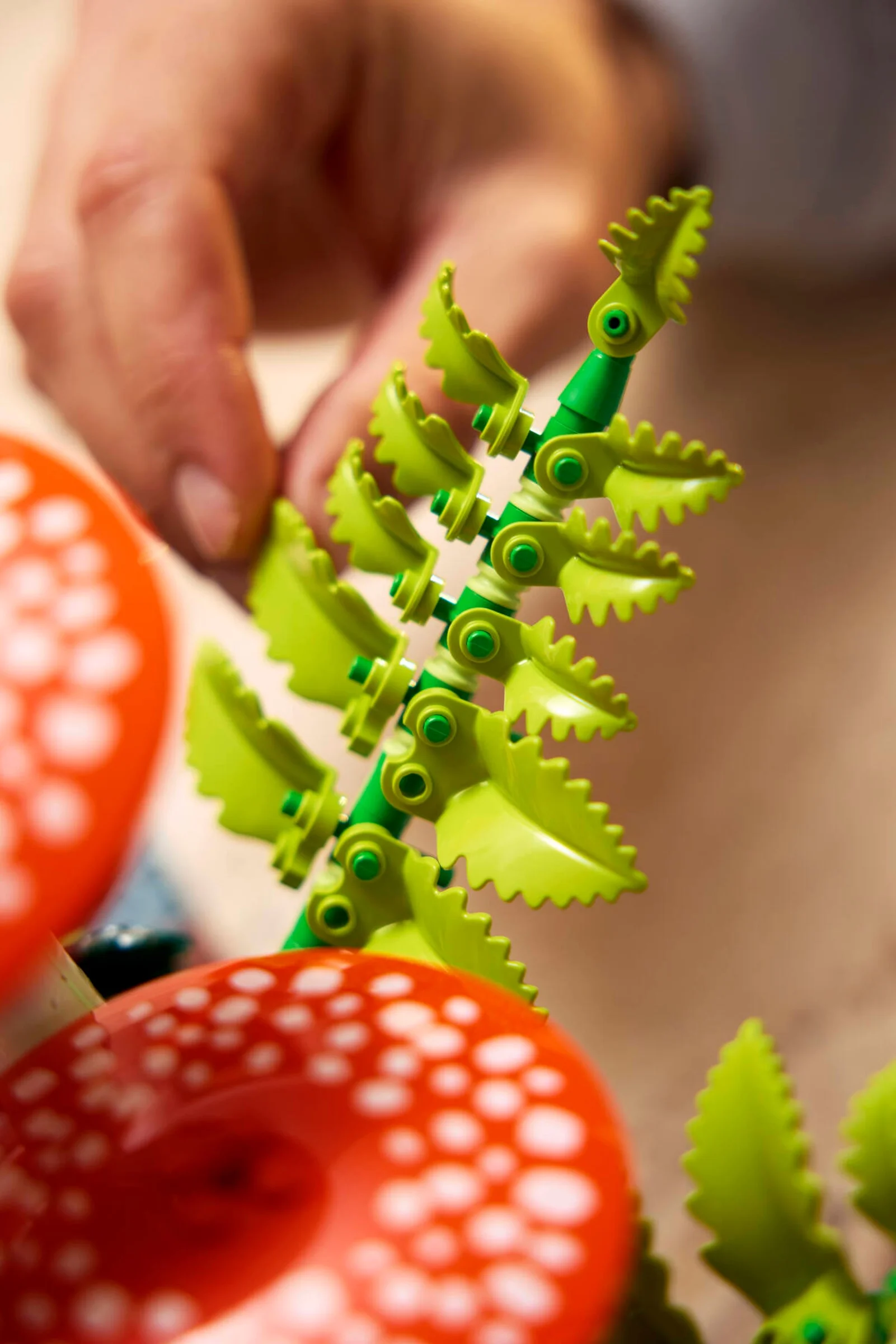 Detaliu cu o persoană construind ciuperci LEGO, arătând o tulpină verde cu frunze zimțate și o pălărie roșie cu pete albe.