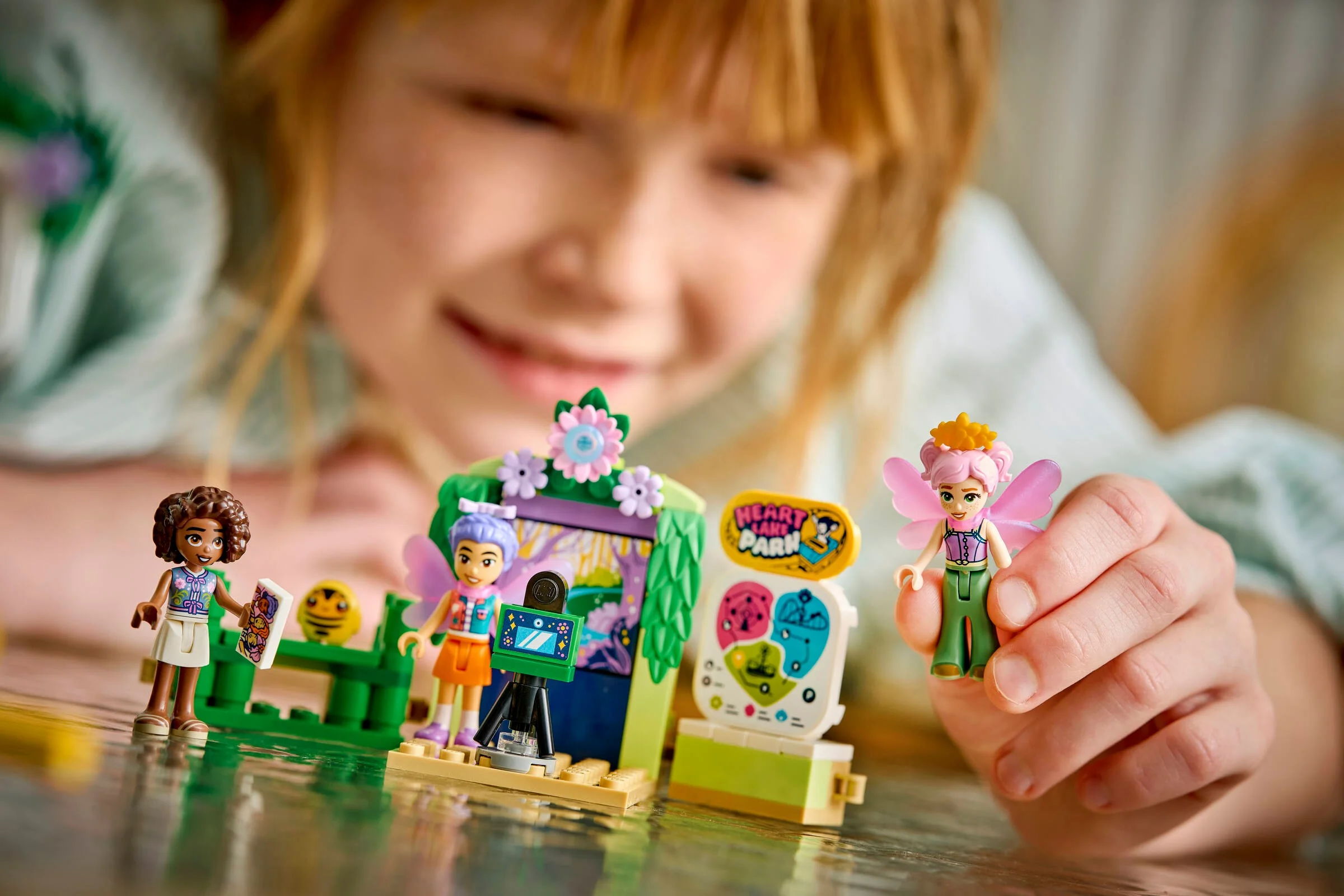 Prim-plan cu minifigurinele LEGO Friends și decorul scenic. Zâmbetul copilului în fundal adaugă căldură imaginii.