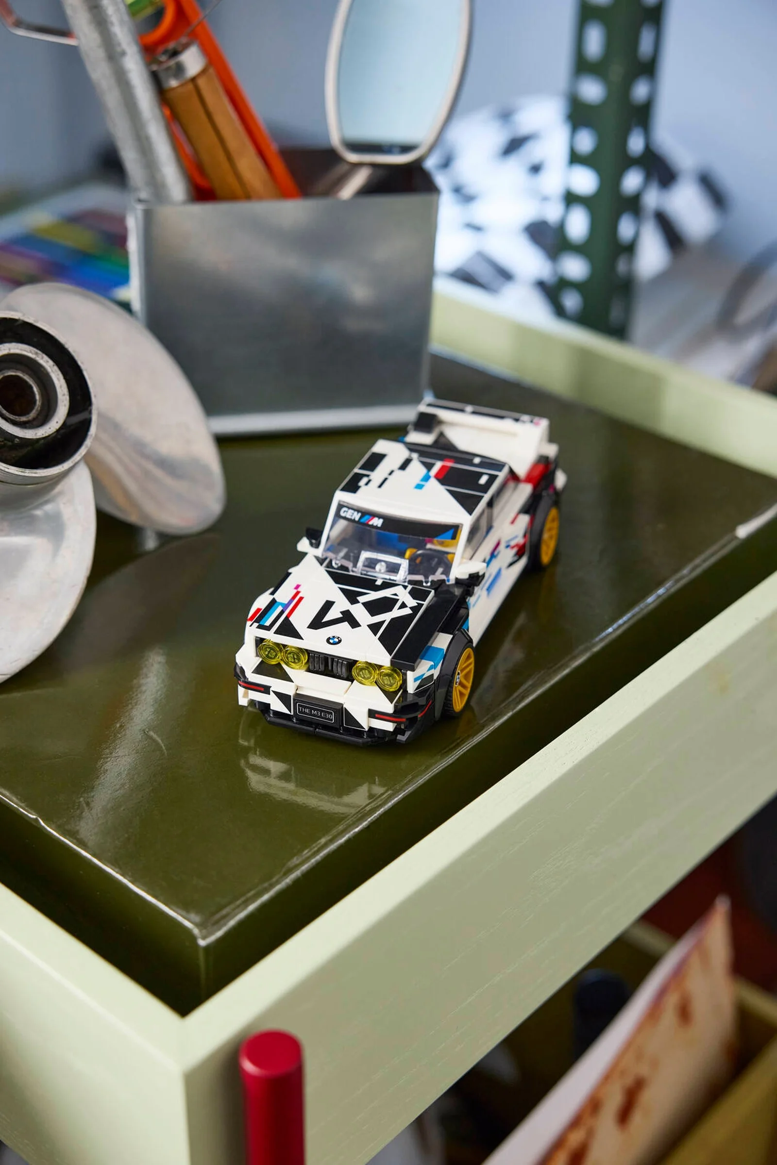 Modelul LEGO BMW M3 (E30) pe o măsuță verde, cu alte obiecte în fundal. Detaliu atrăgător.