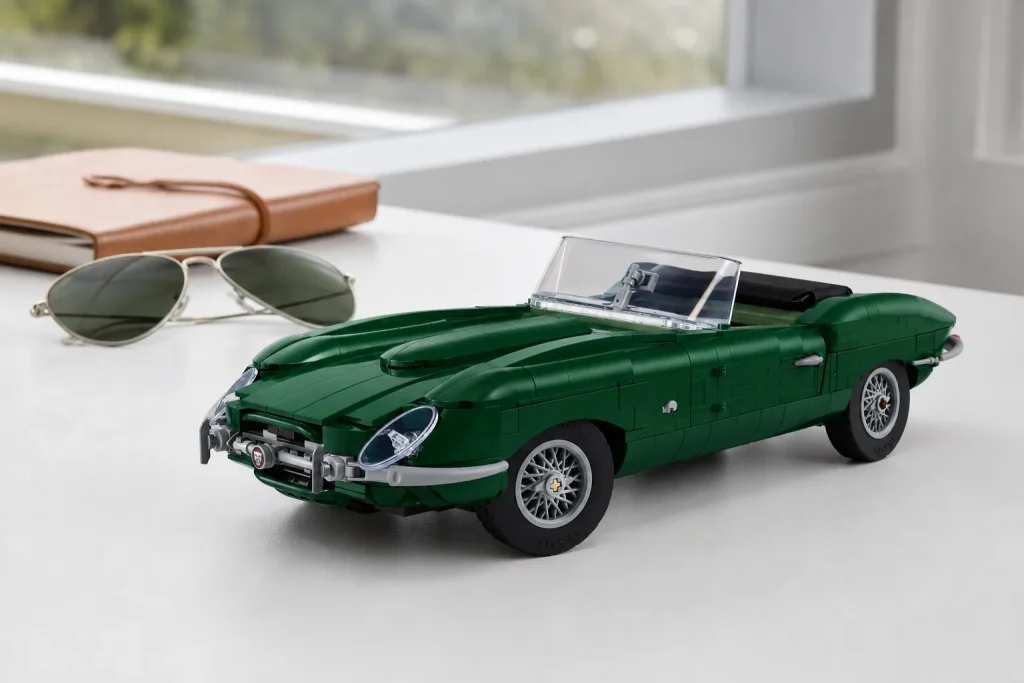 Modelul LEGO Icons 11381 Jaguar E-Type este prezentat dintr-un unghi jos pe un birou alb minimalist, sub o lumină naturală difuză, alături de ochelari de soare clasici.
