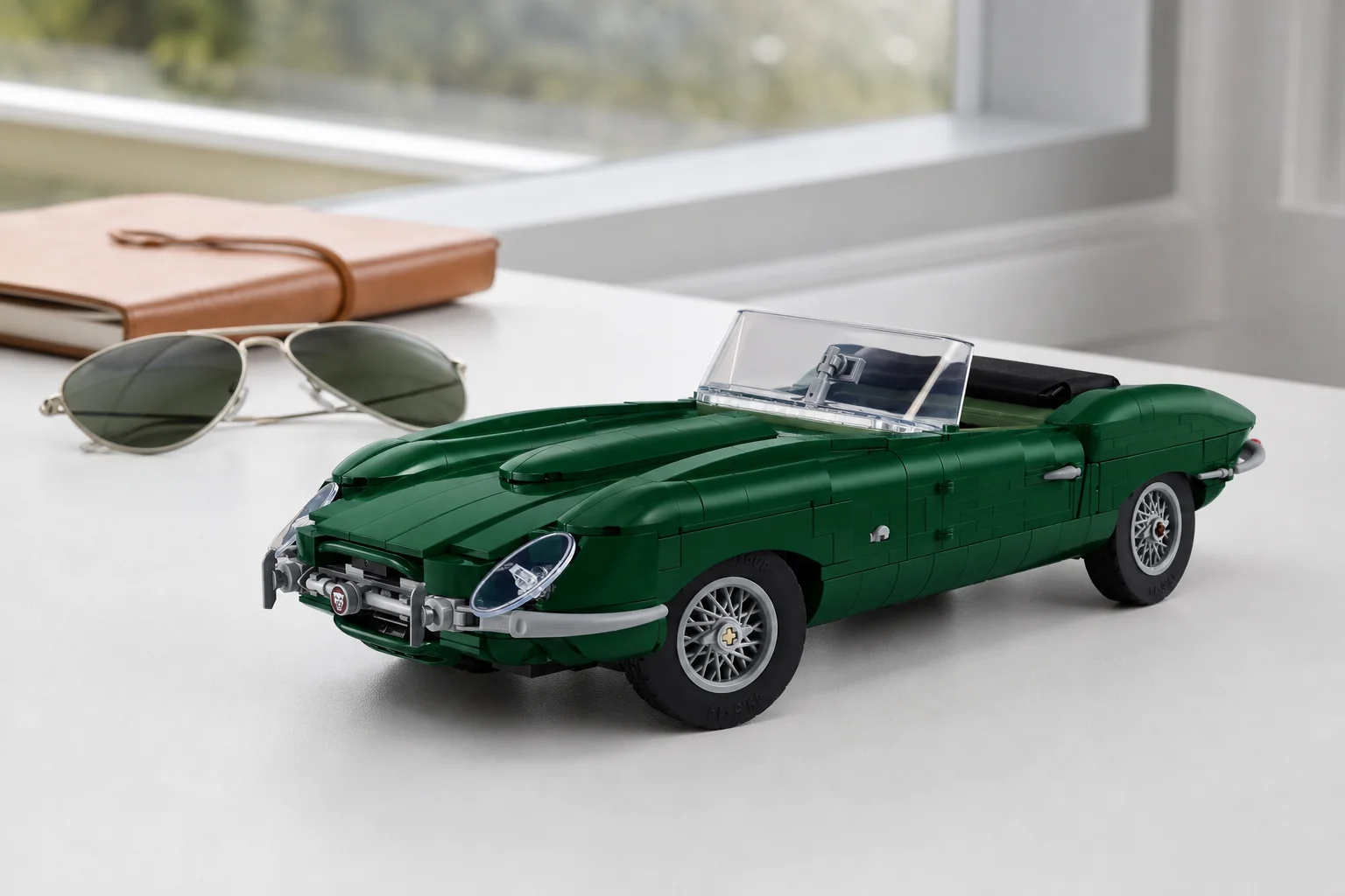 Modelul LEGO Icons 11381 Jaguar E-Type este prezentat dintr-un unghi jos pe un birou alb minimalist, sub o lumină naturală difuză, alături de ochelari de soare clasici.