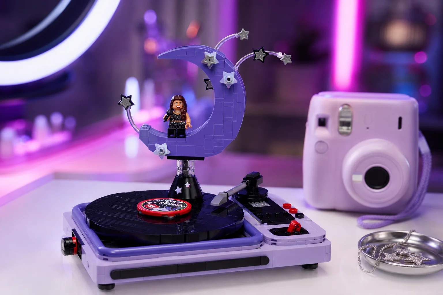 Setul LEGO 43029 Luna de concert a Oliviei Rodrigo expus pe o masă de toaletă albă, strălucitoare, decorată cu un aparat foto instant lila și luminată de o bandă LED în nuanțe de violet și roz.